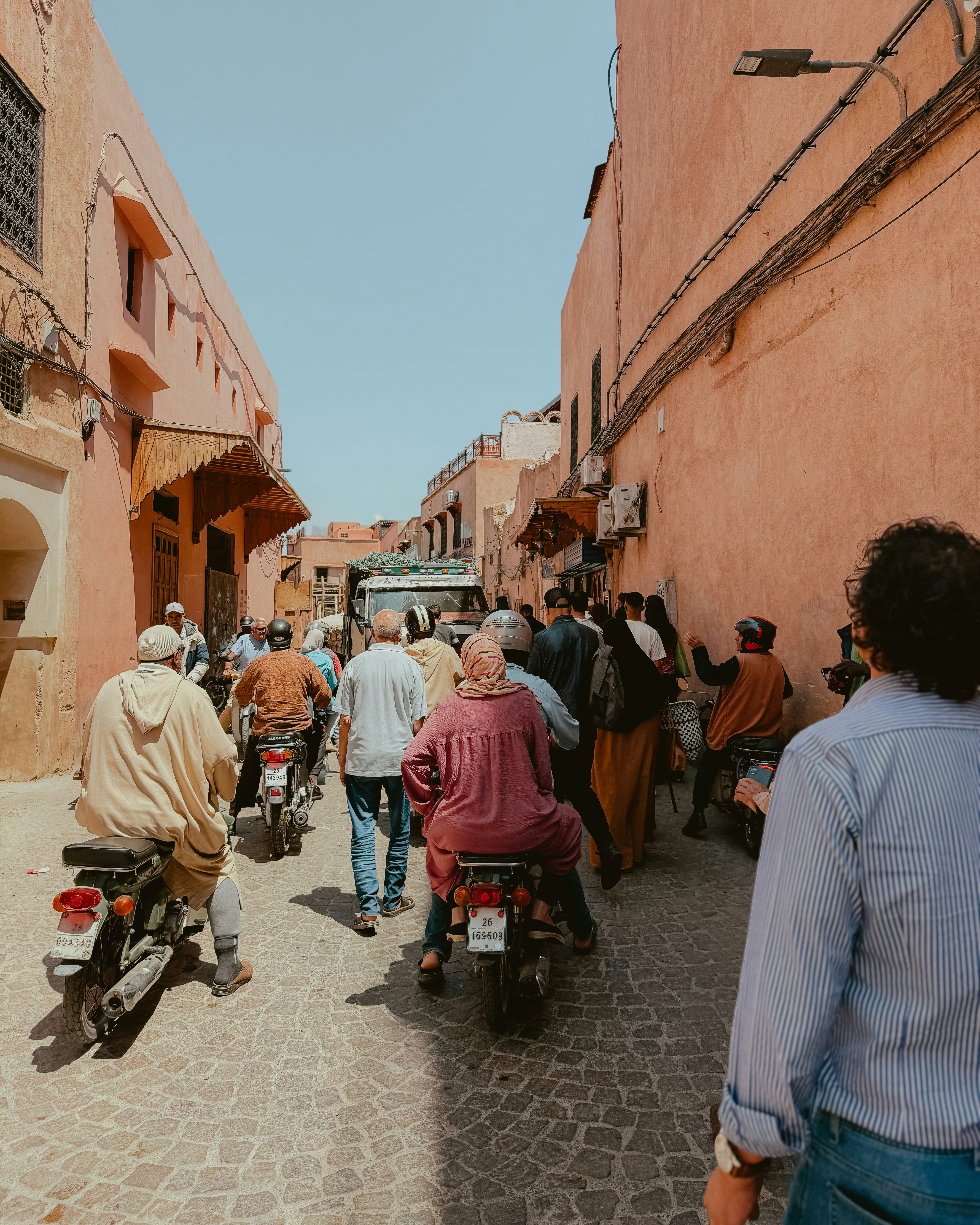 Motorcycles ride down a narrow street with buildings.