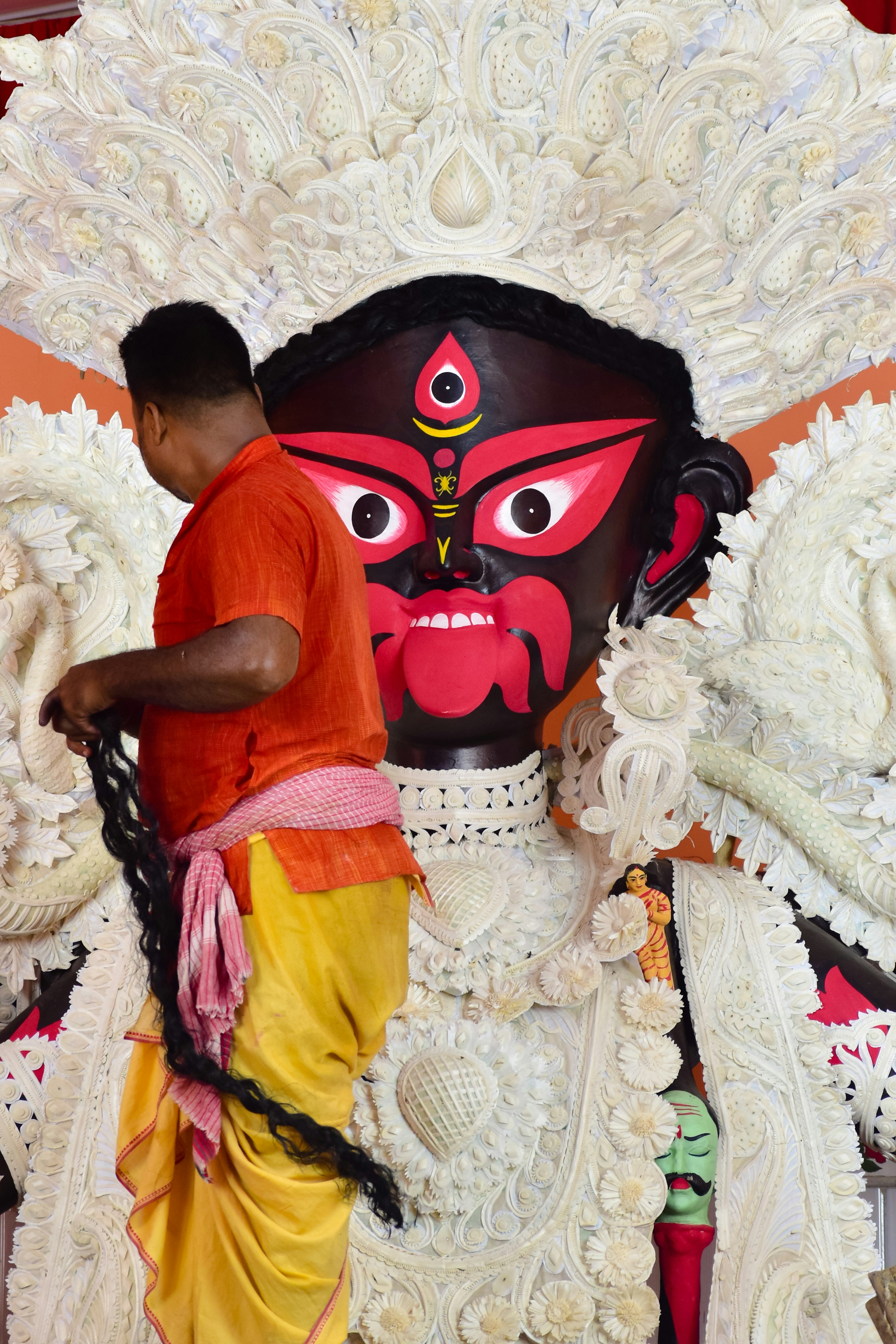 A man with a long braid in orange and yellow stands in front of a massive white-carved deity with a vivid red face and ornate headdress.