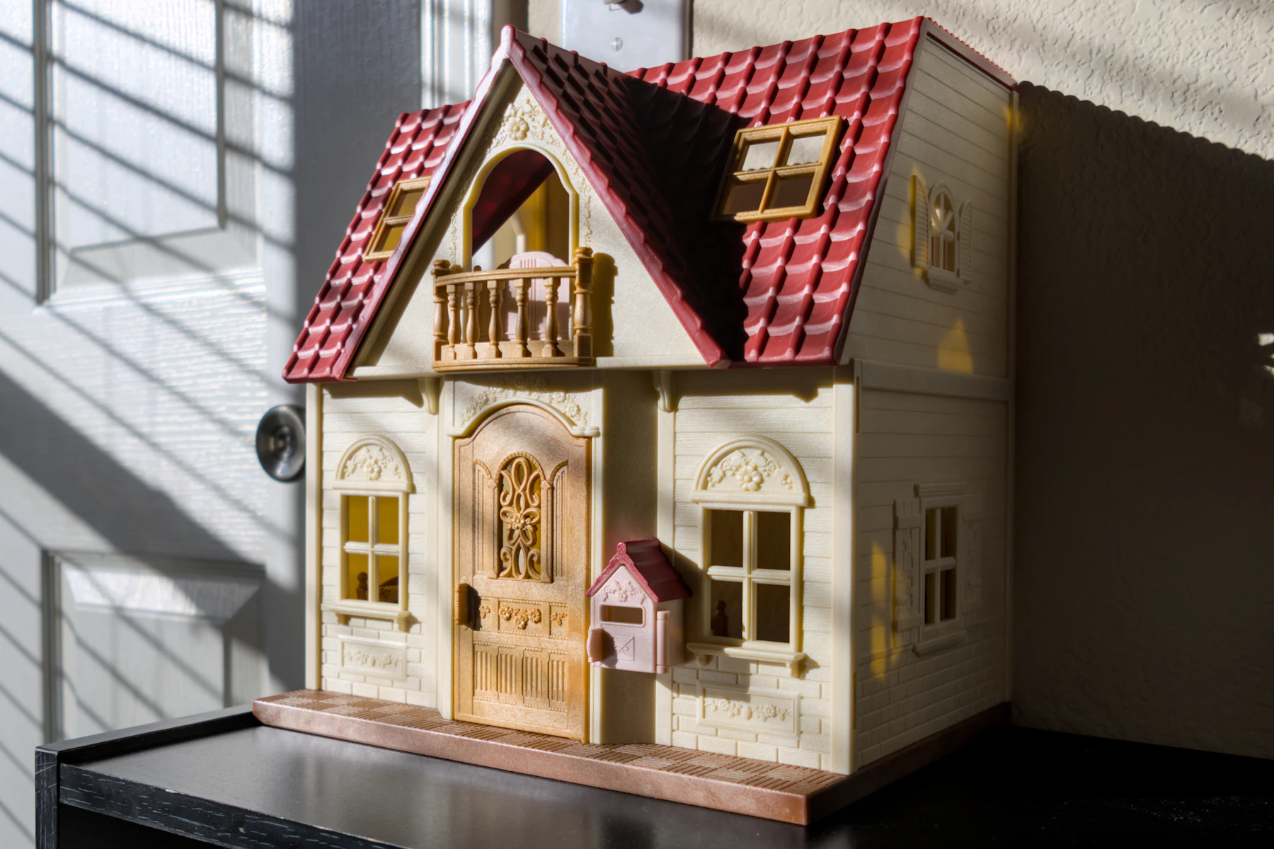 A cream dollhouse with a red tiled roof, a balcony and an arched front door in window light
