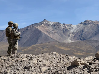 Two soldiers stand on a rocky outcrop overlooking mountains.