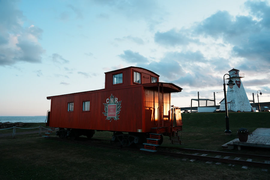 A red CNR railway caboose converted to accommodation sitting beside a white Maritime lighthouse at dusk, the ocean and sky visible behind in shades of orange and blue