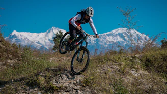 Mountain biker jumps over a dirt mound with snow-capped mountains.