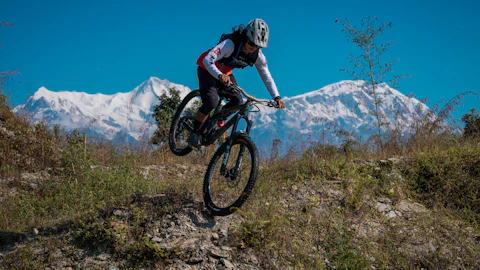 Mountain biker jumps over a dirt mound with snow-capped mountains.