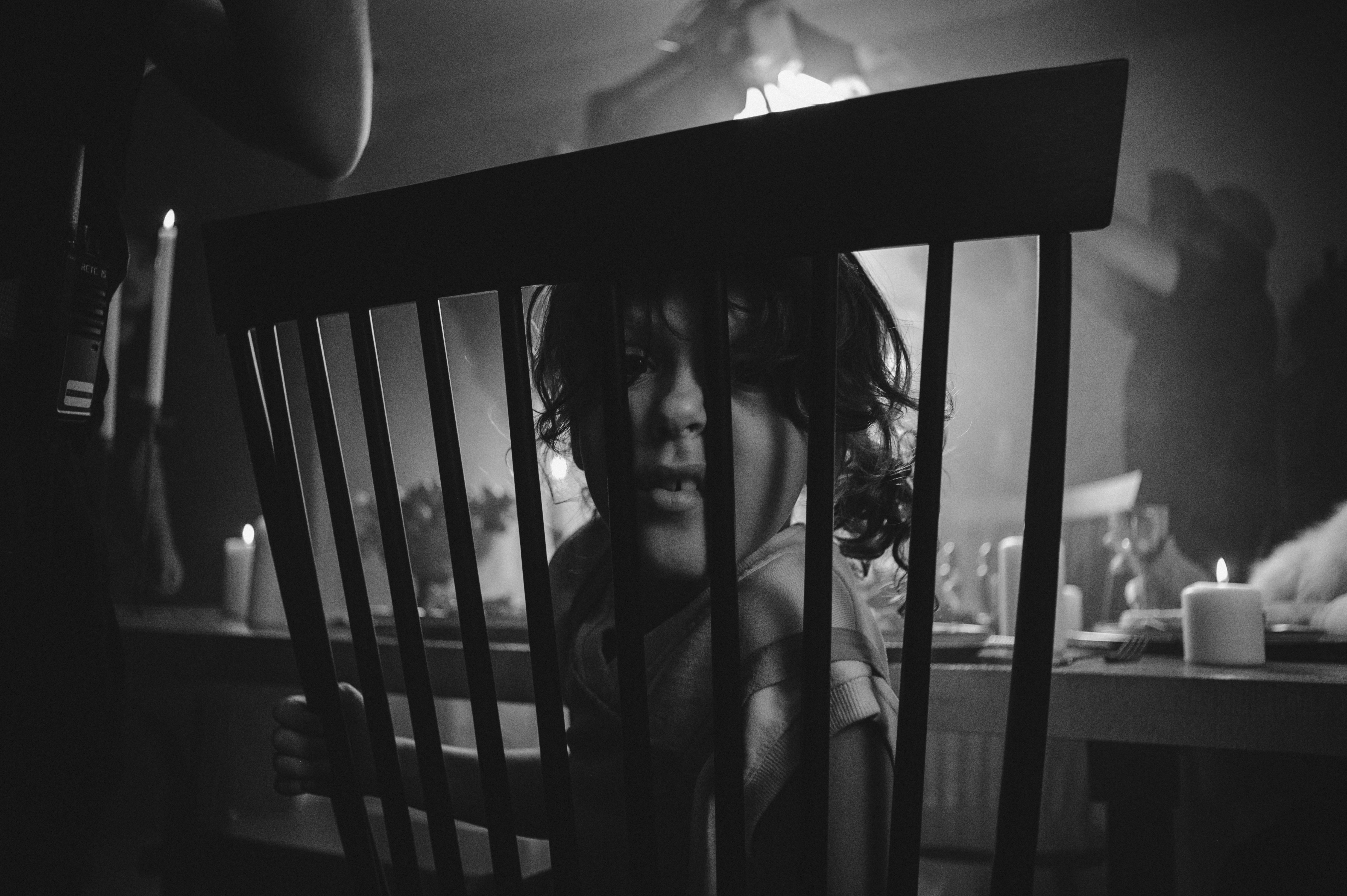 Child peeking through chair slats in dimly lit room.