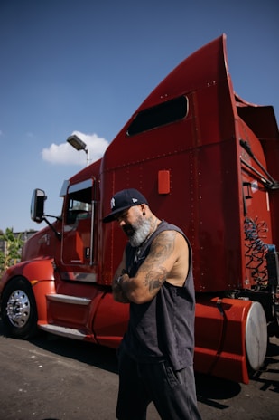 Man with tattoos stands by a red semi-truck.