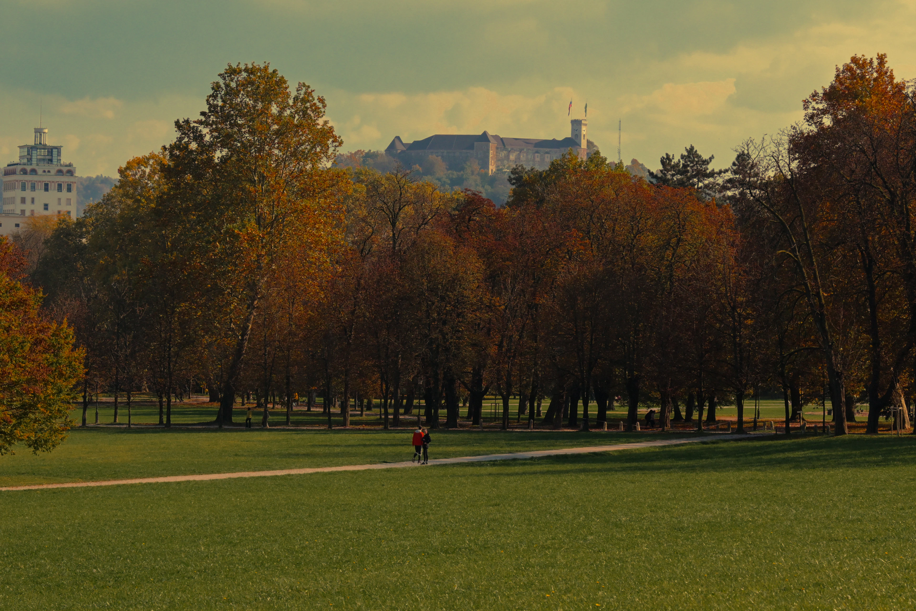 An autumn afternoon on a wide park lawn in Ljubljana, two walkers on the gravel path beneath russet and gold trees, with Ljubljana Castle on its wooded ridge in the distance