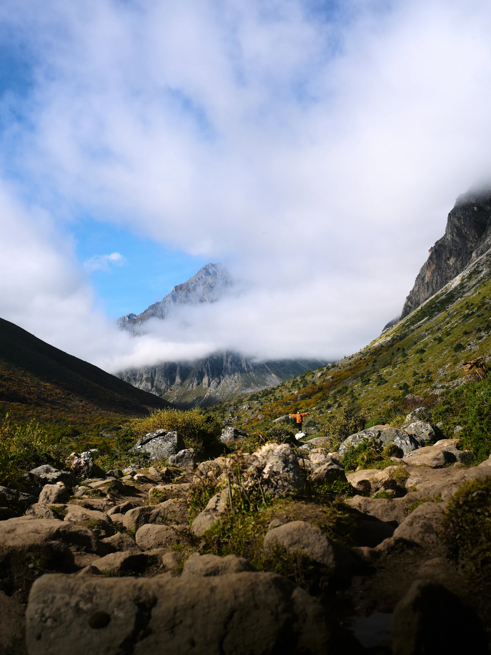 Photograph of A lone hiker in an orange jacket below a rocky peak disappearing into thick cloud.