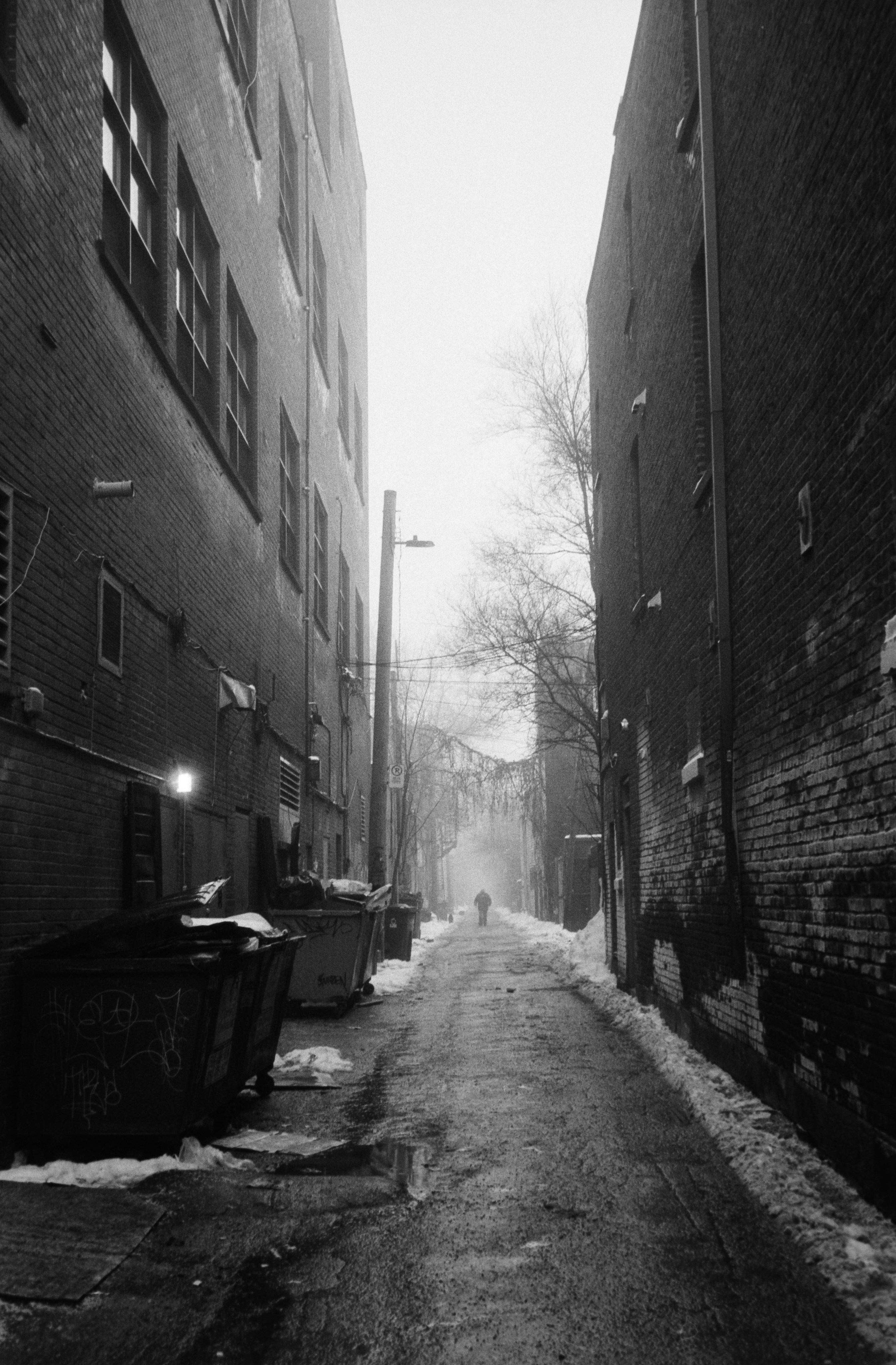 Une silhouette solitaire marche dans une ruelle urbaine enneigée.