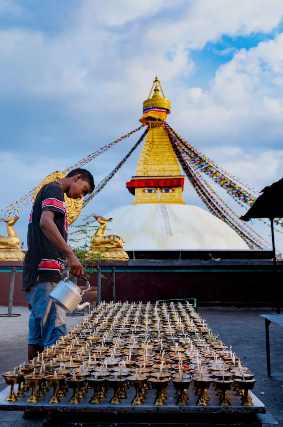 A man tending a tray of brass butter lamps on the Boudhanath kora, the white dome rising behind