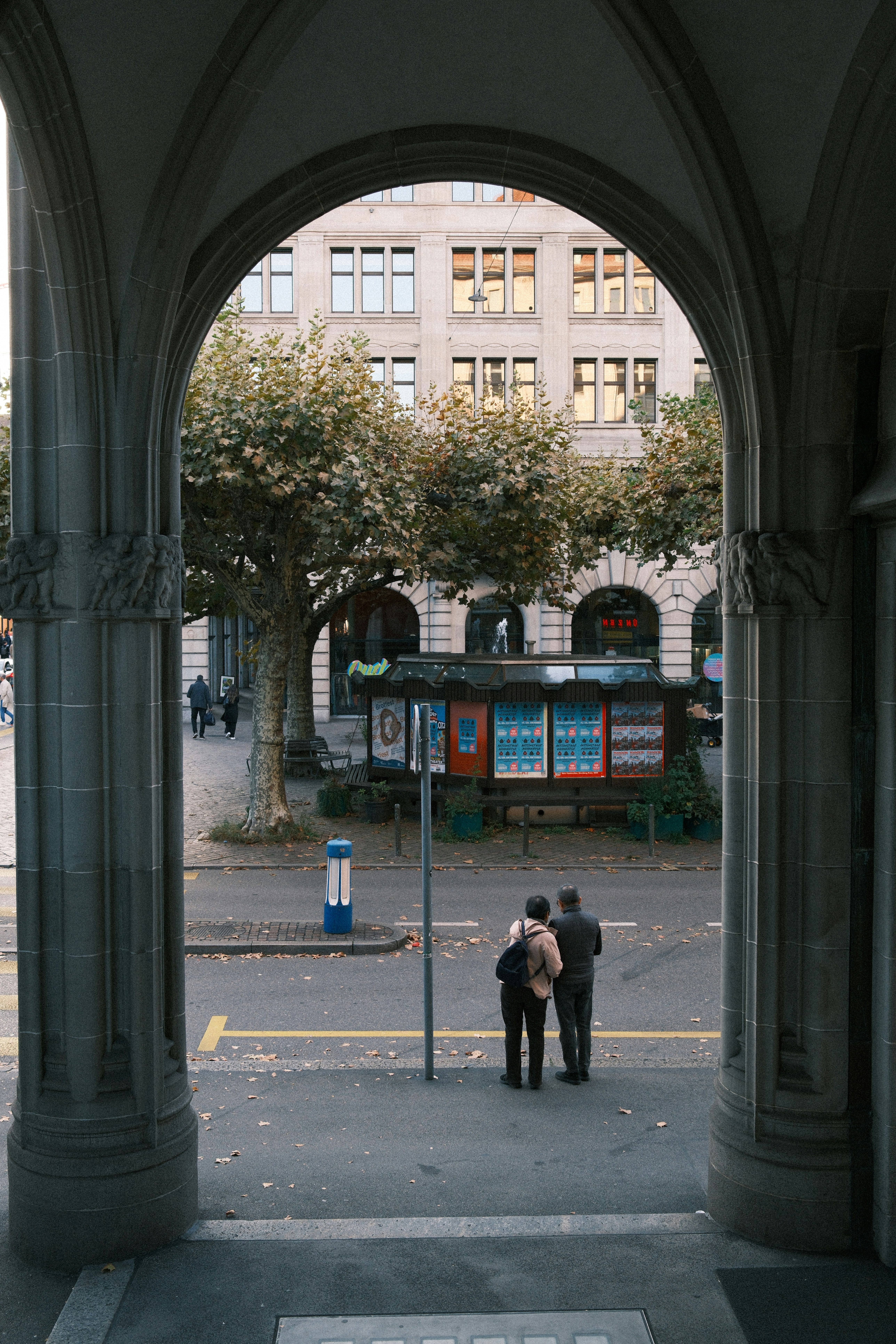 Two people stand under an archway, looking at a street.