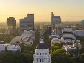 The white dome of the California State Capitol rising above trees at sunrise, with Sacramento's skyline behind it.