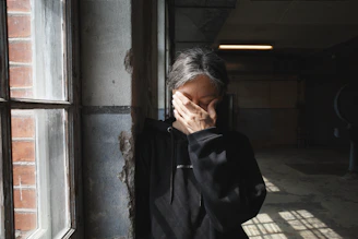 Person covers face with hand near window.