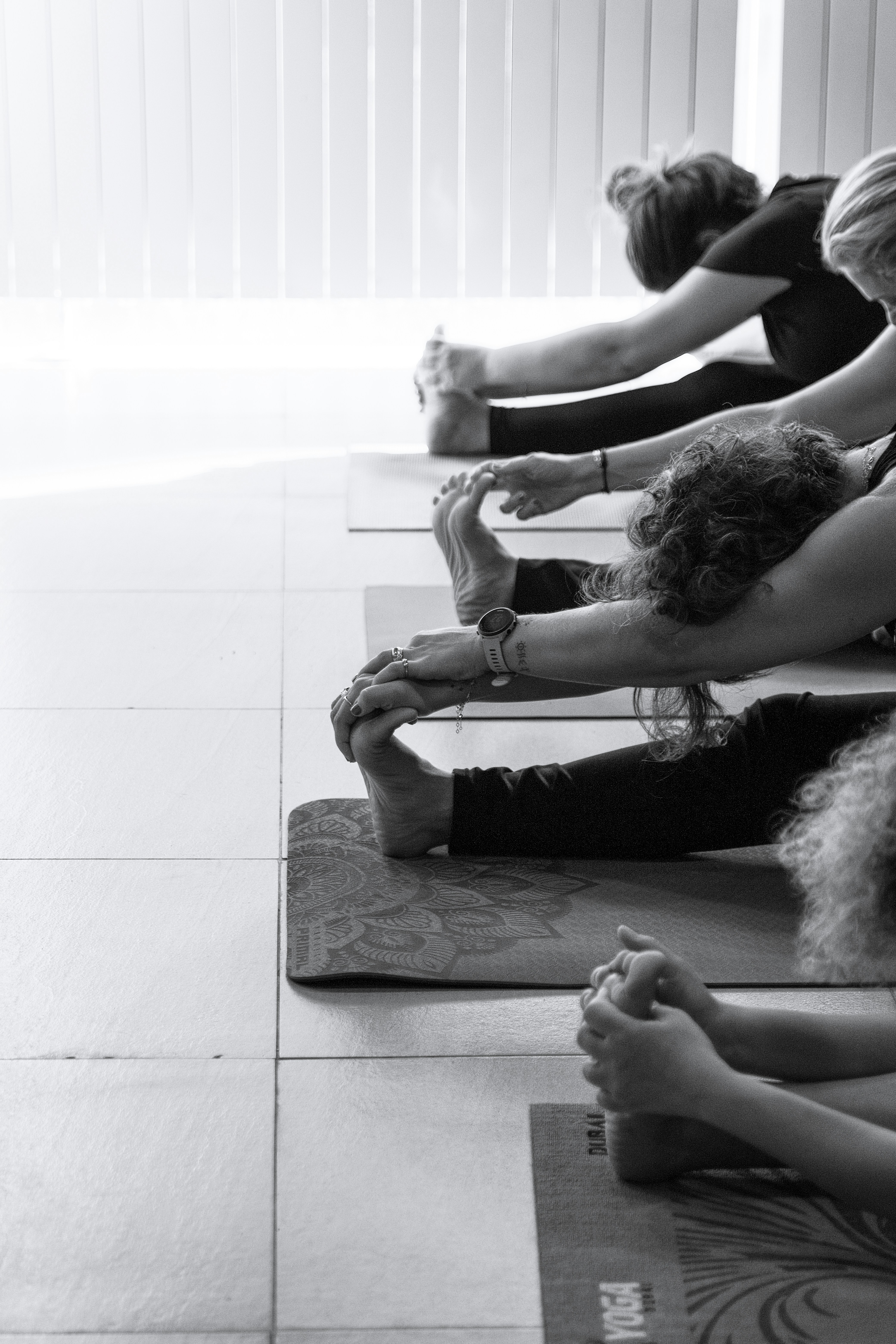 students in a yoga class sat on floor reaching forward to grab foot of straight leg