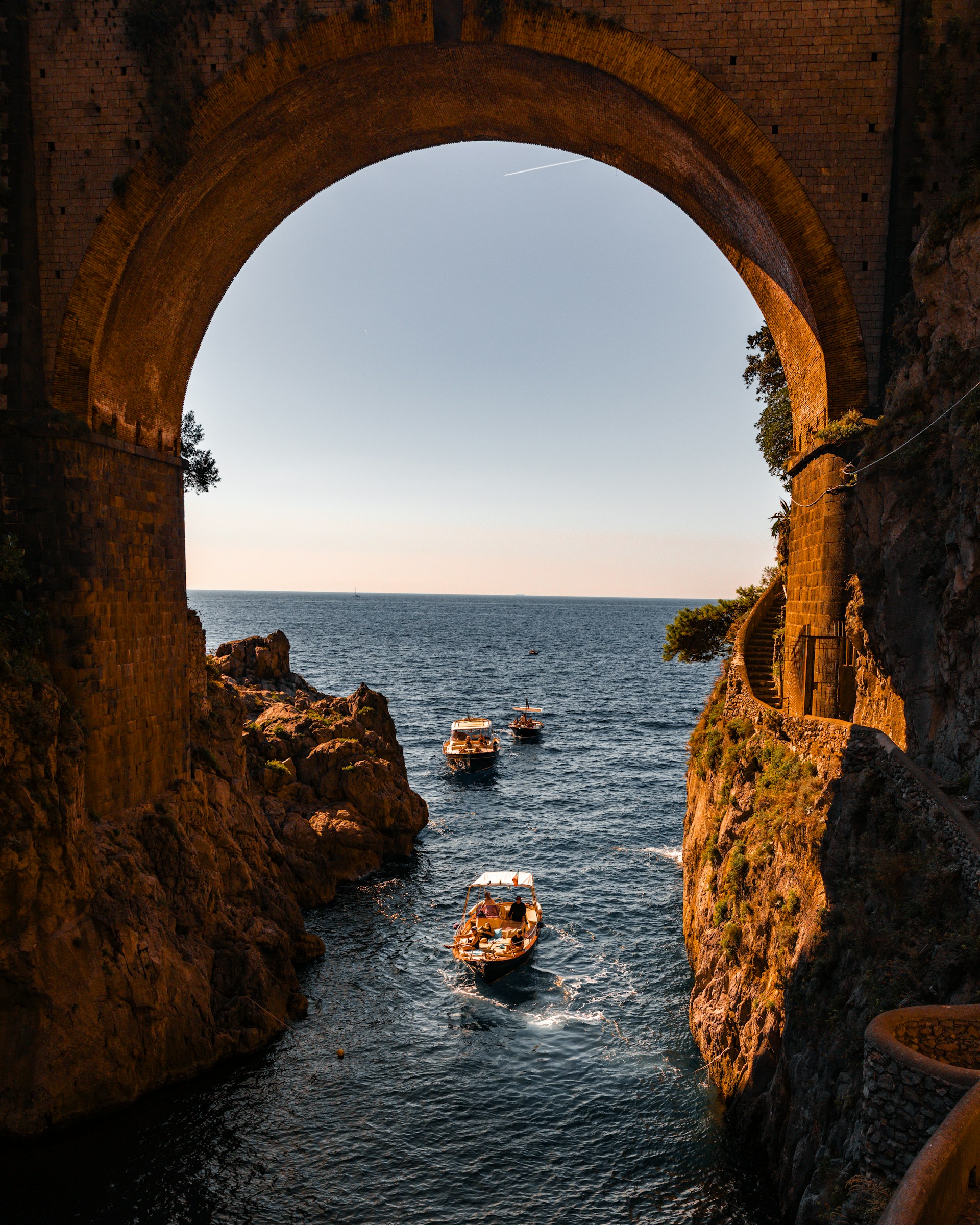 Boats sail through a narrow sea channel under an arch.