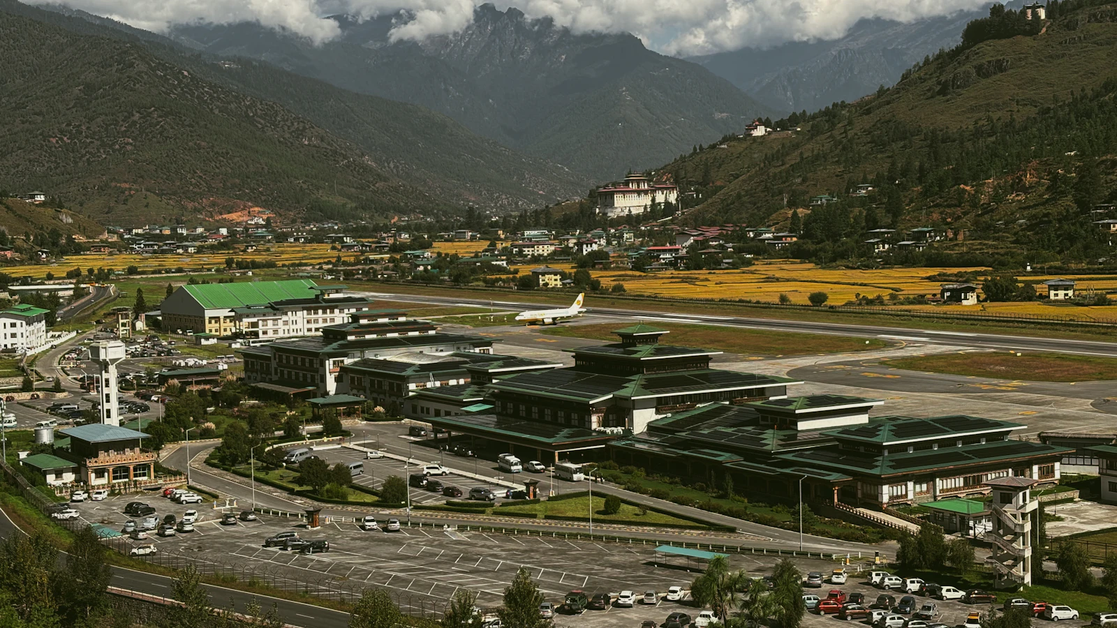 Paro airport's terminal buildings and runway on the valley floor, pine ridges and cloud topped peaks rising behind