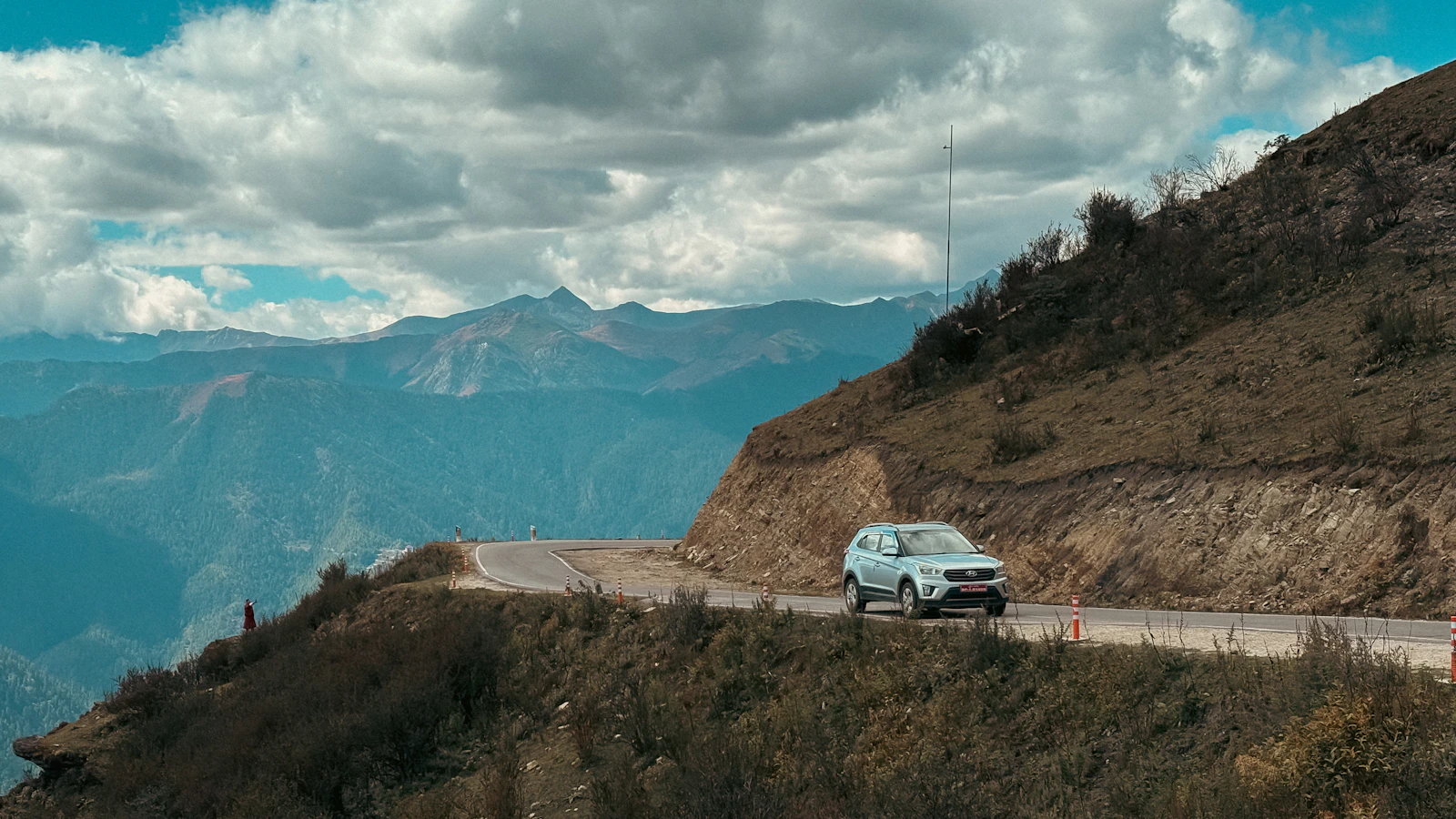 A white SUV rounding a hairpin on a mountain road above Paro, Bhutan, with layered blue ridges and valleys falling away beyond