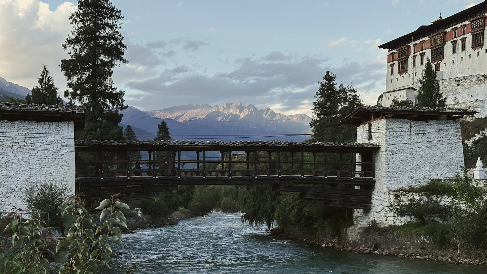 The covered cantilever bridge over the Paro Chhu at dusk, Rinpung Dzong's walls above and snow peaks in the distance