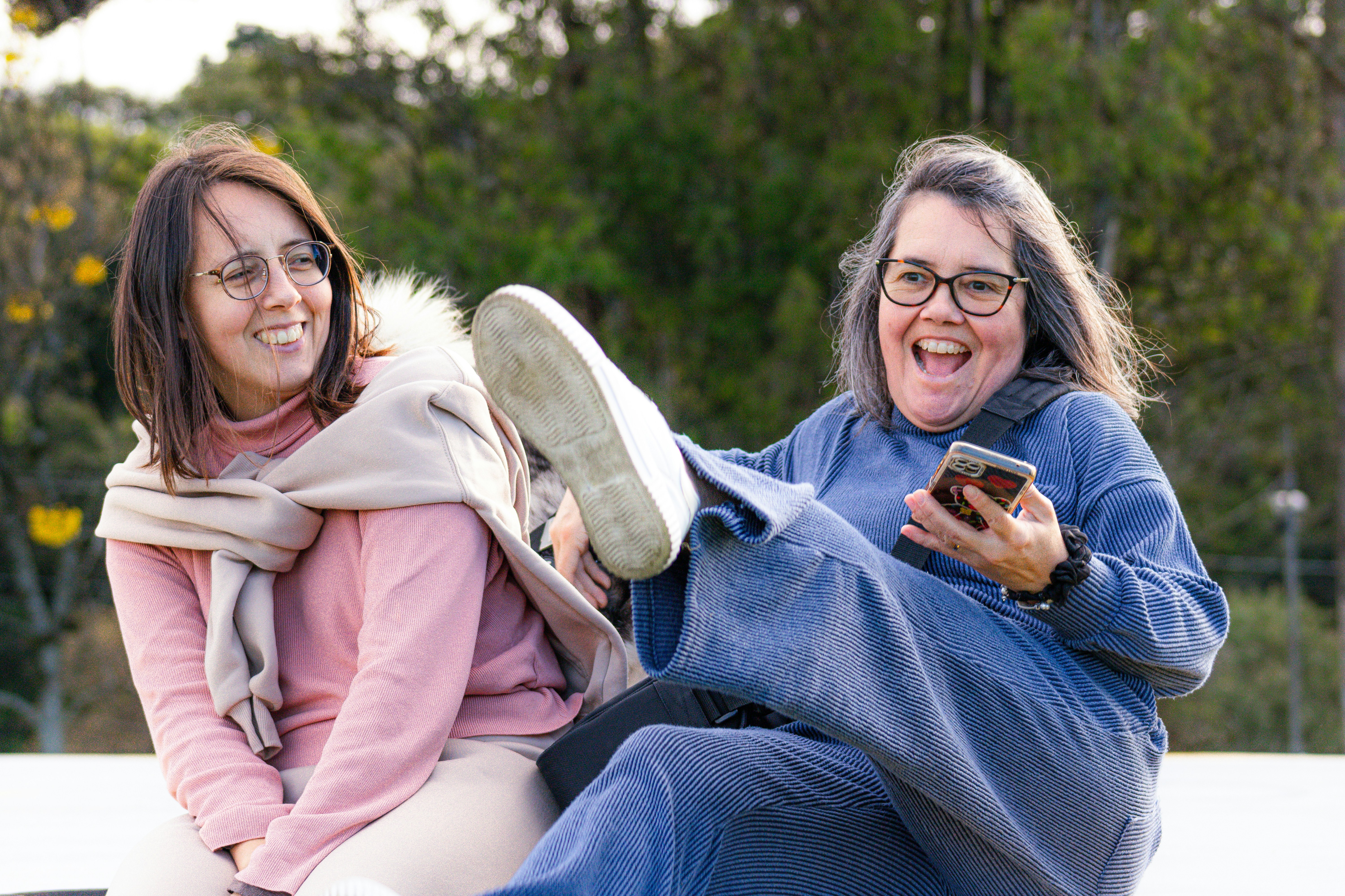 Two women laughing outdoors with one kicking their leg up.