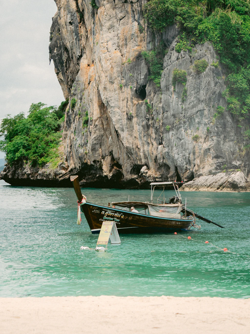 A wooden longtail boat in turquoise water beneath a limestone cliff in Krabi