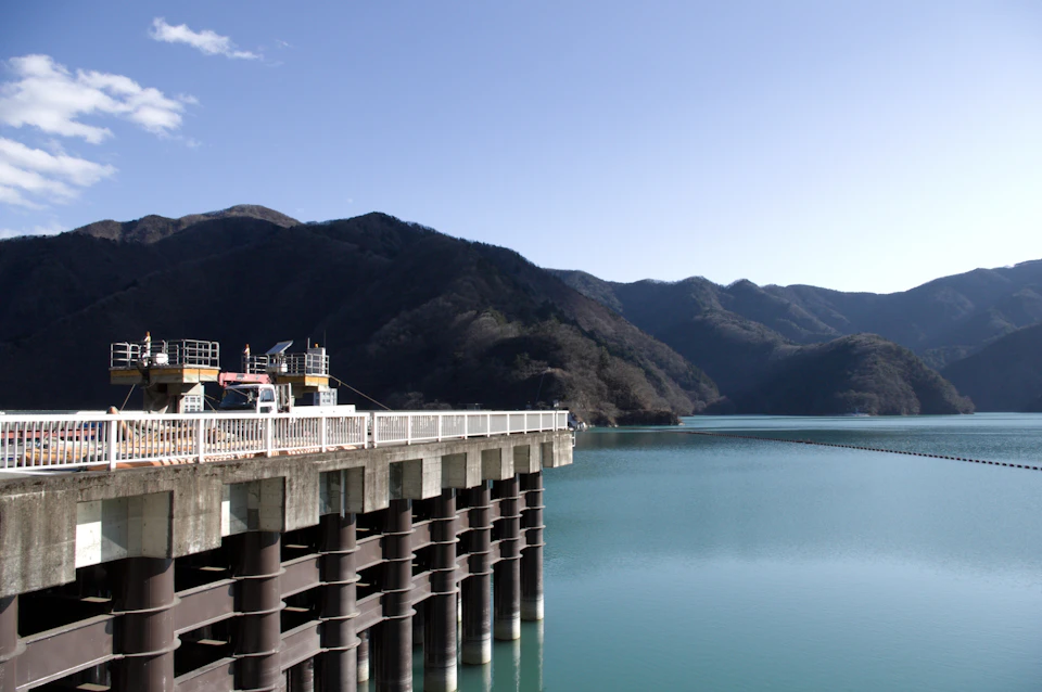 Two people stand on a concrete pier extending into a serene lake surrounded by hills