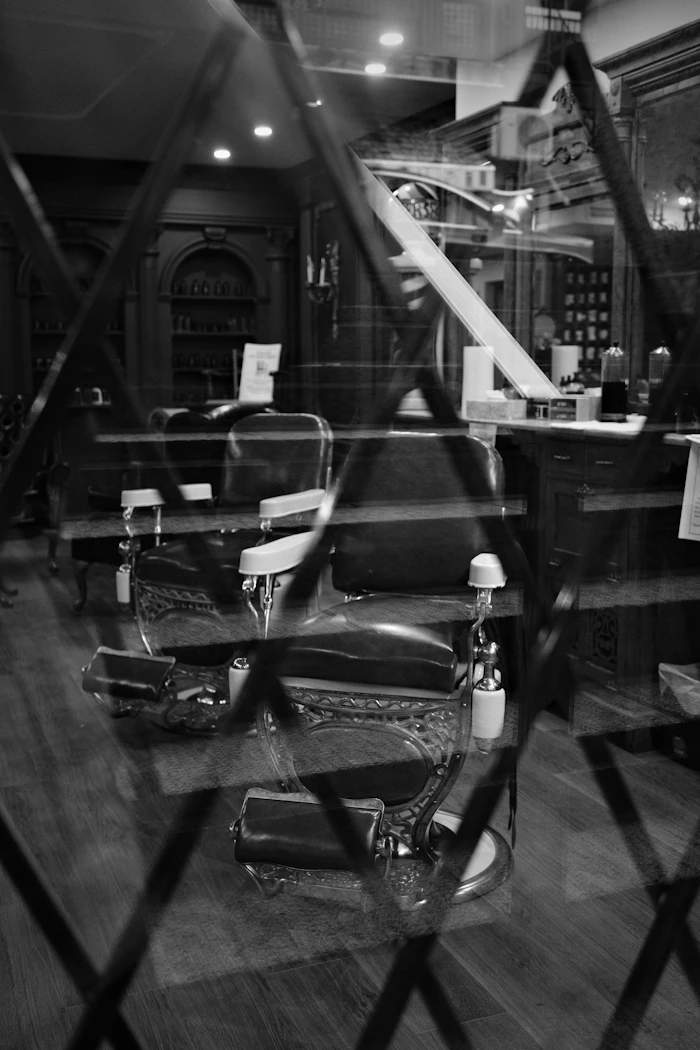 Black and white view of vintage barber chairs seen through a shop window with a lattice reflection.