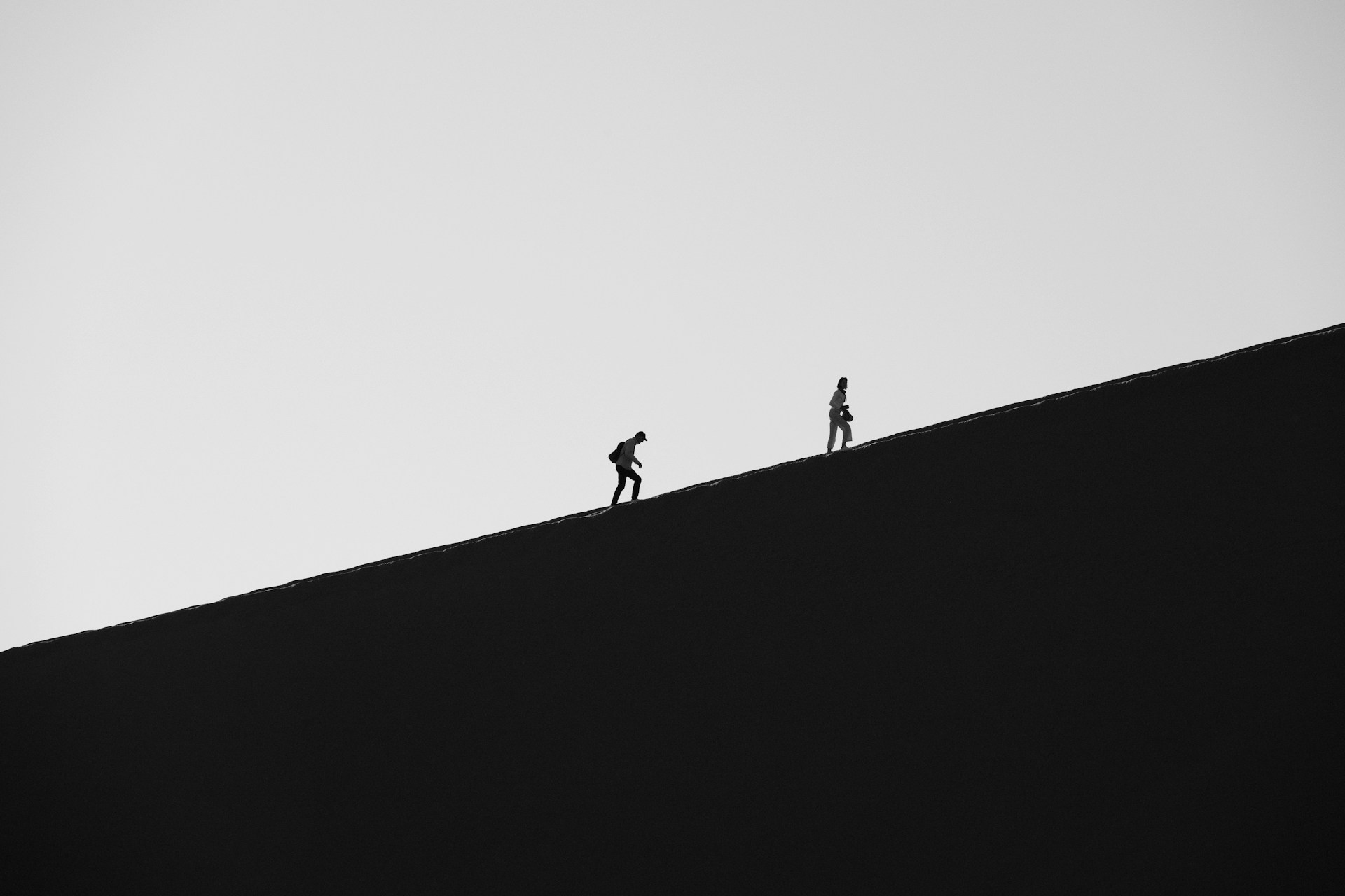 Two hikers ascend a dark, barren dune.