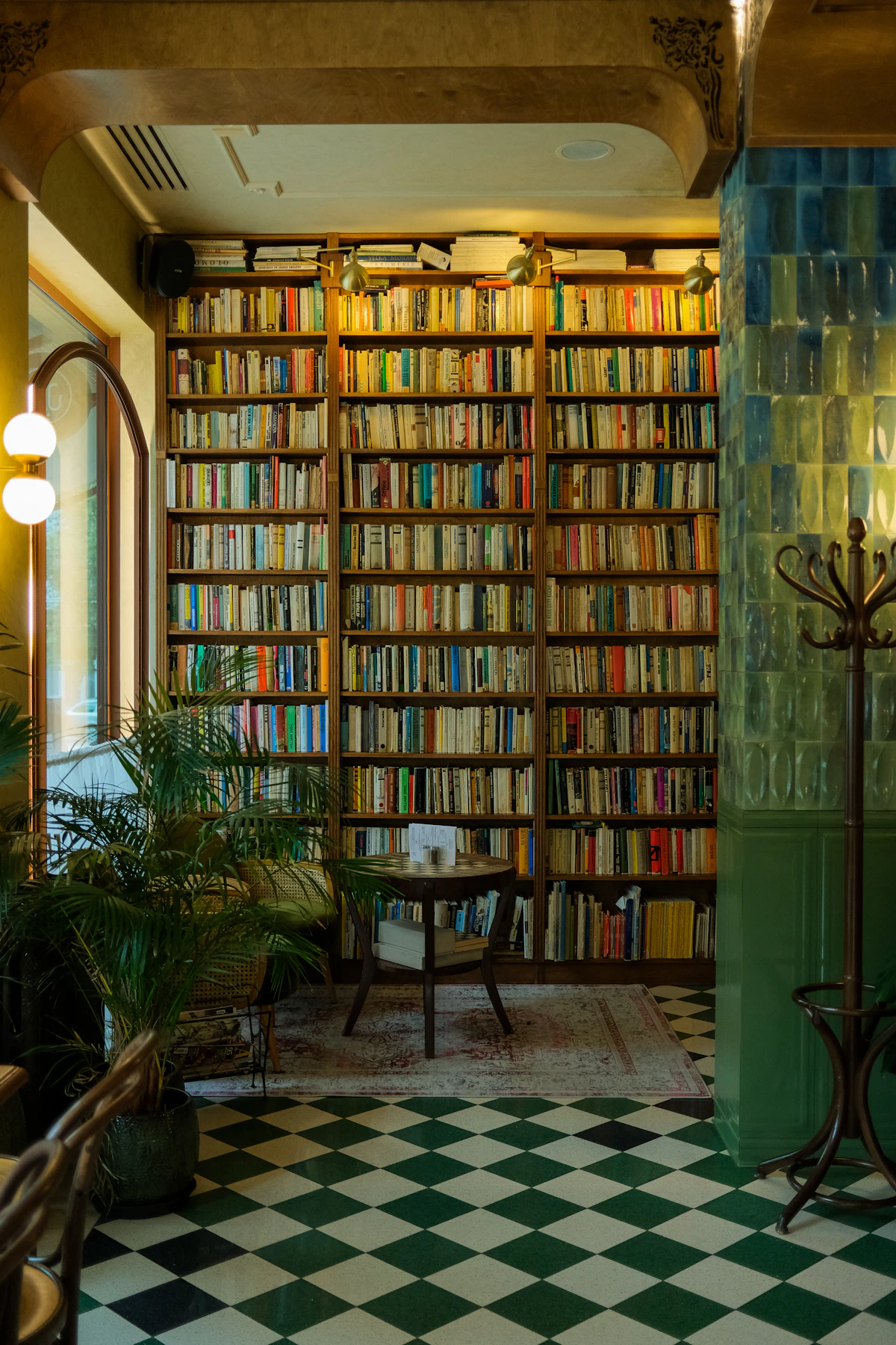A warm library of colourful books with green tiles, palms and an arched window
