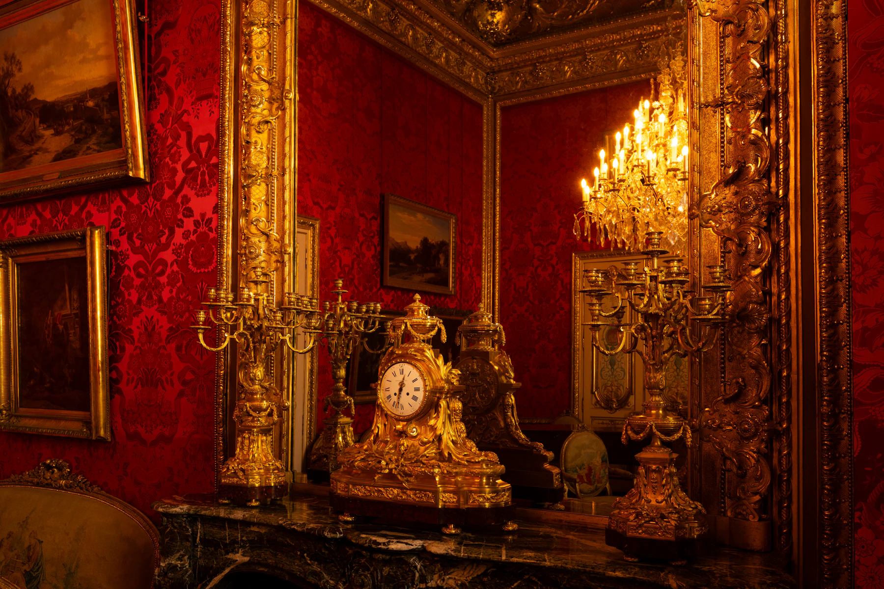 A gilt mantel clock standing between candelabra on a marble mantelpiece in a red room