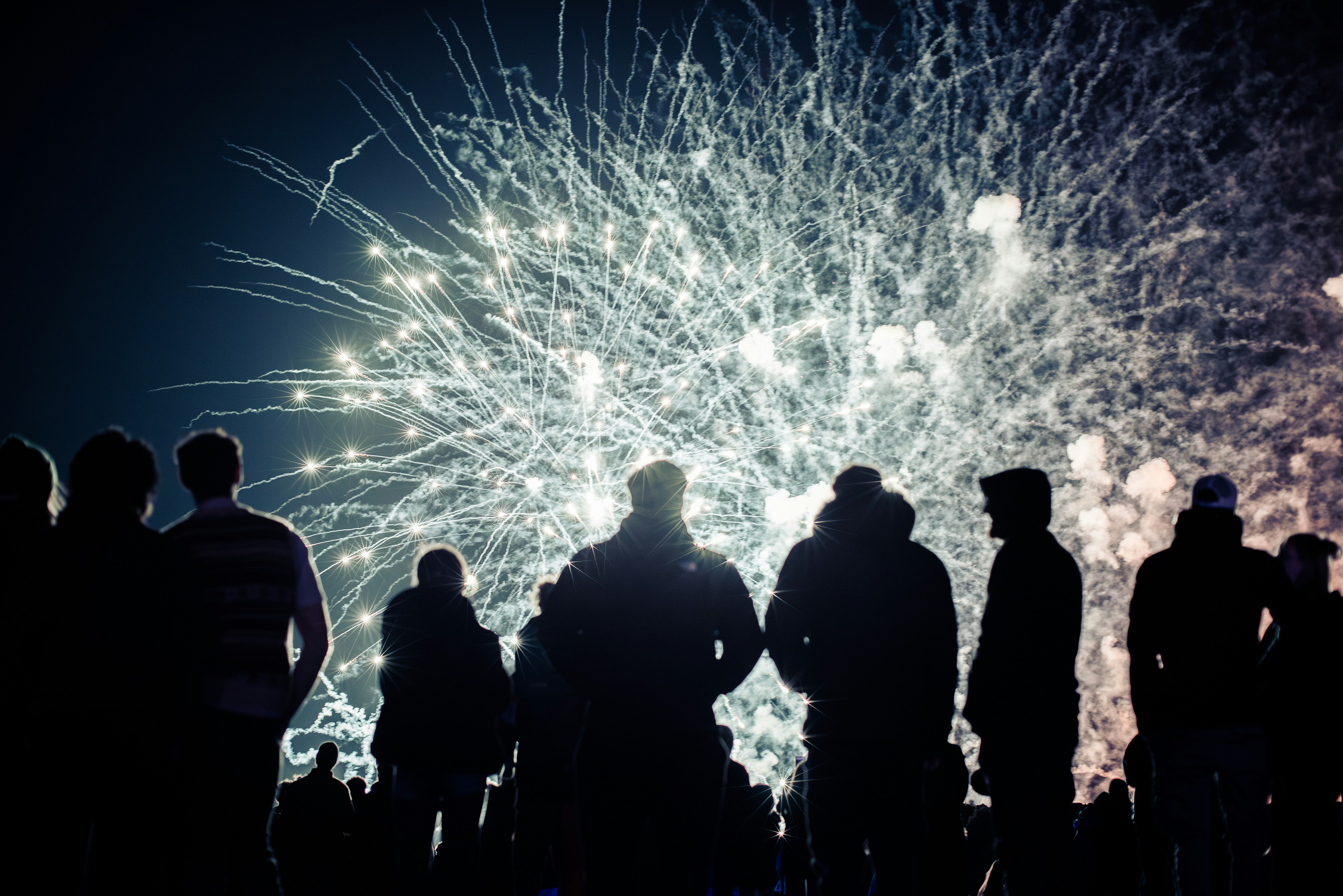 People stand in silhouette before a massive burst of white fireworks, the radiant light and drifting smoke turning the night sky into a glowing canvas.