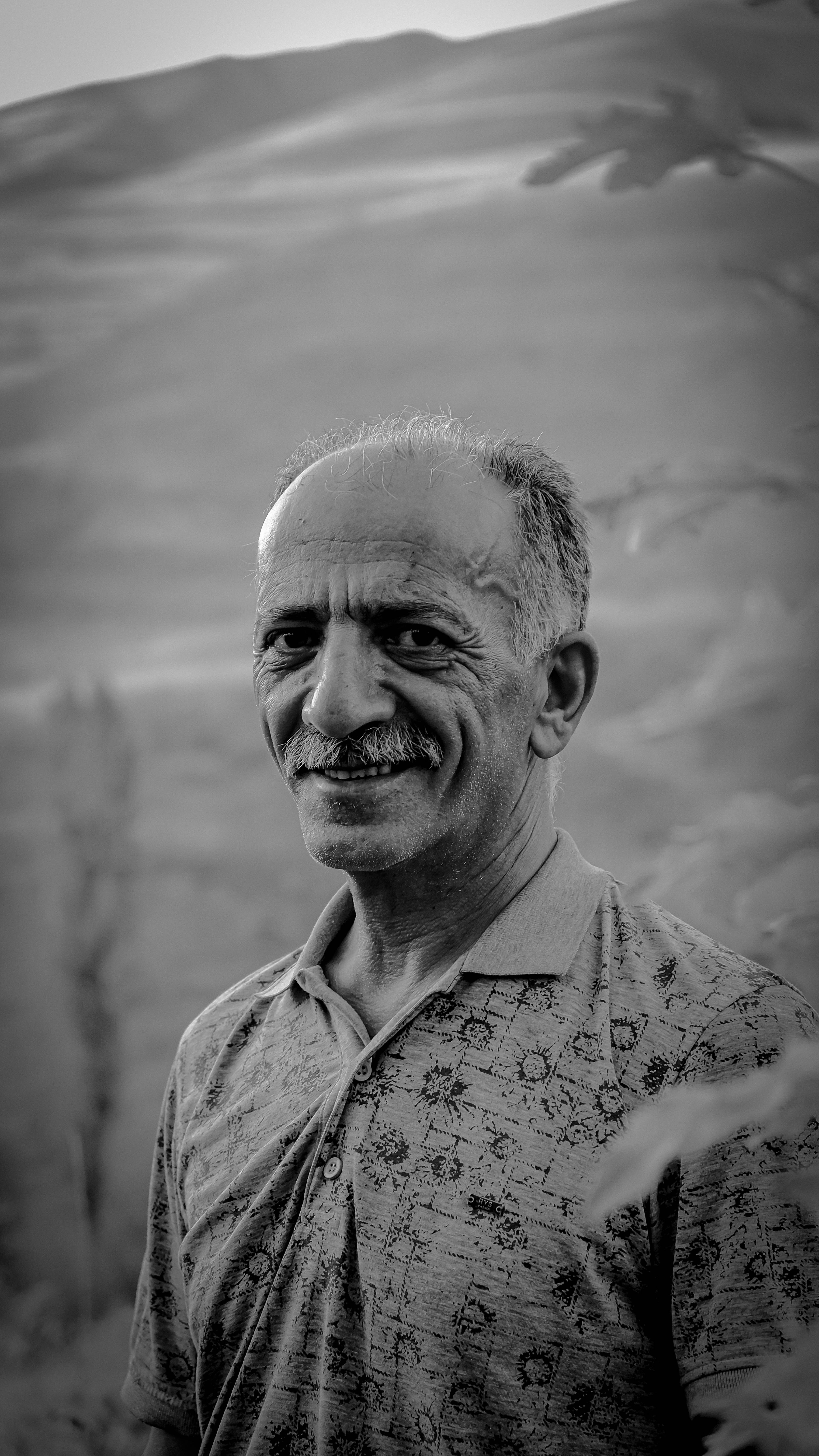 Man with mustache smiles in black and white outdoor setting.