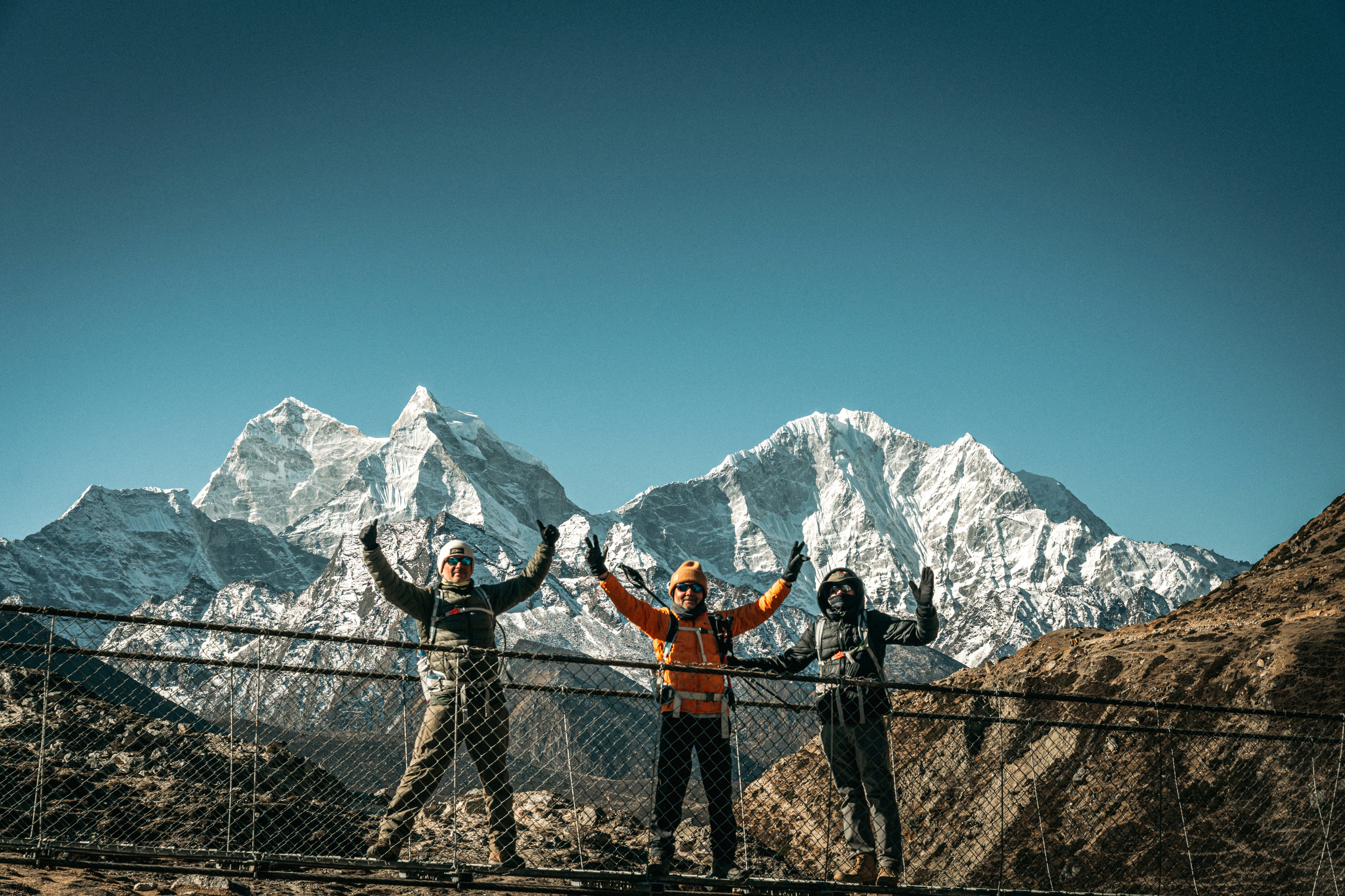Three hikers celebrate on a suspension bridge with mountains.
