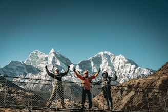 Three hikers celebrate on a suspension bridge with mountains.