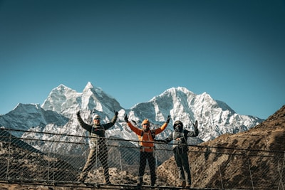 Three hikers celebrate on a suspension bridge with mountains.