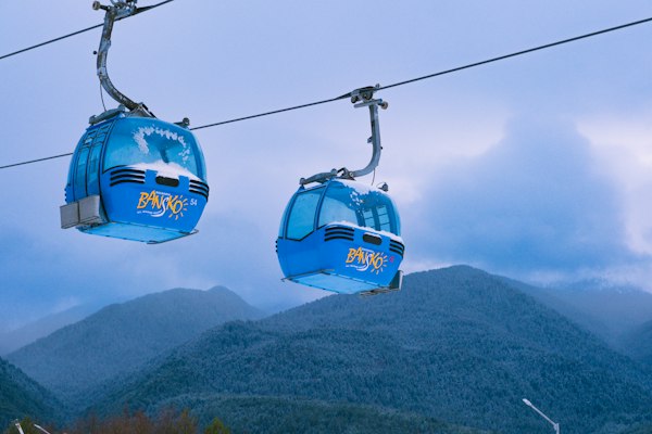 Two blue Bansko gondola cabins with BANSKO lettering on the side, travelling above a forested Pirin ridge