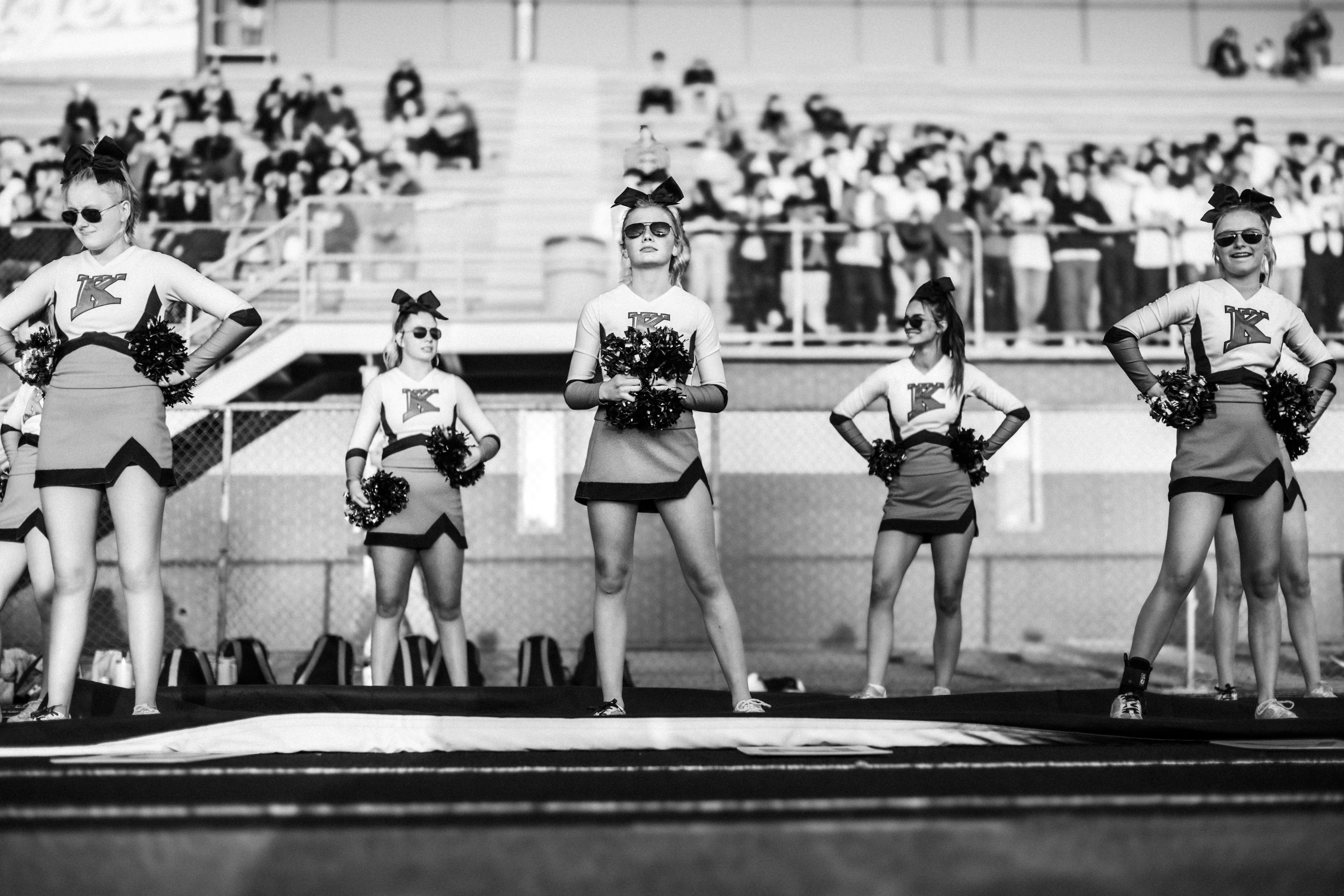 Five cheerleaders pose on a field with spectators behind them.