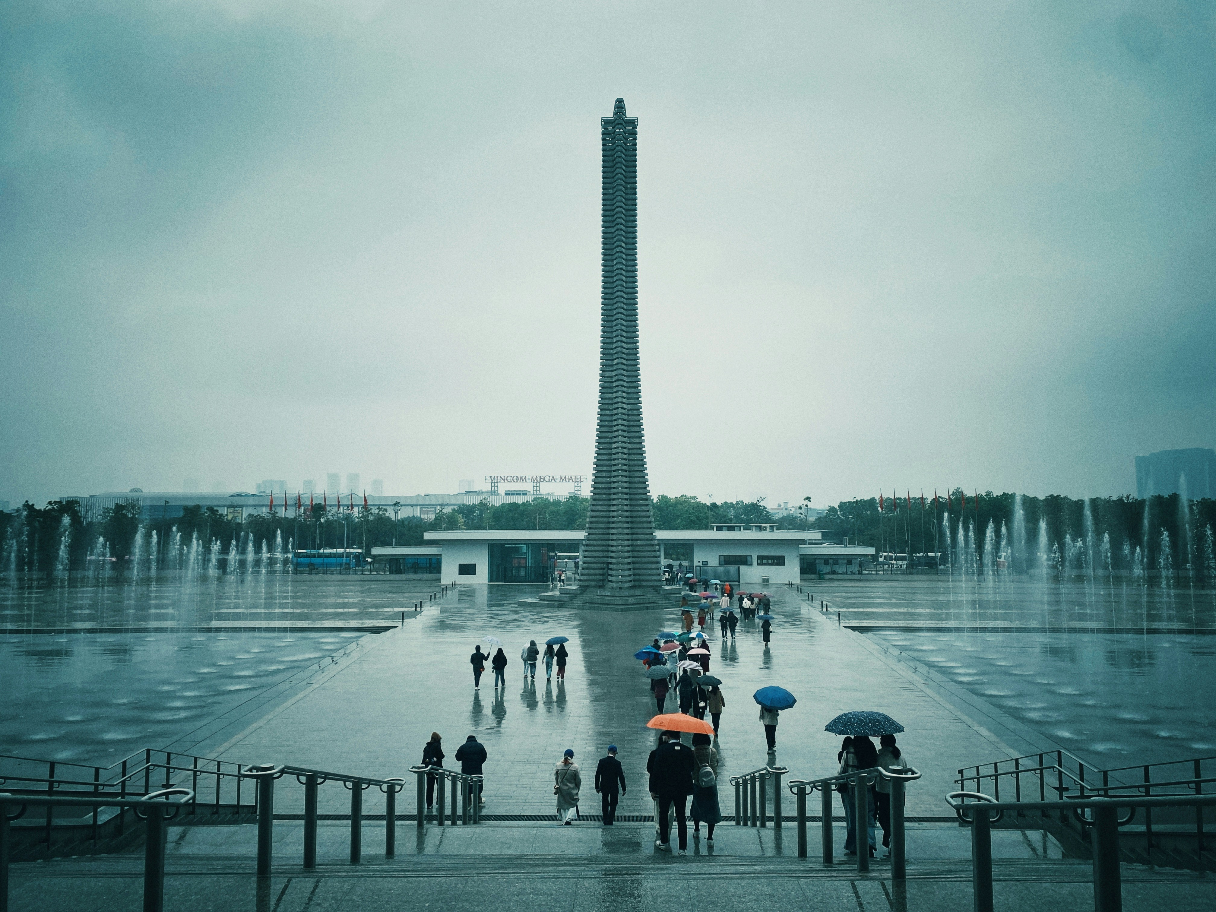 People with umbrellas walk through a plaza with a monument.