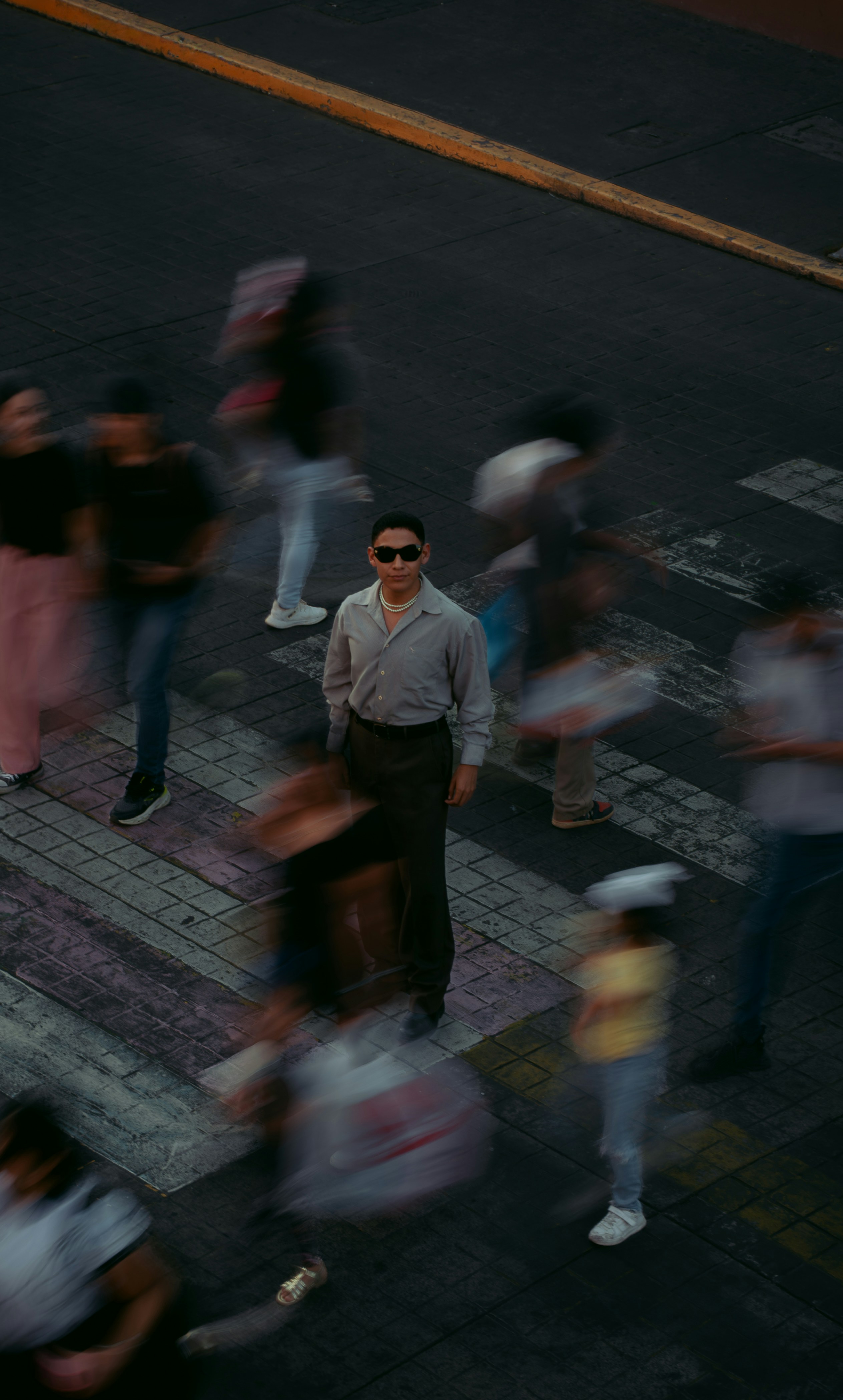 Man in sunglasses stands still on crosswalk amidst blurred people.