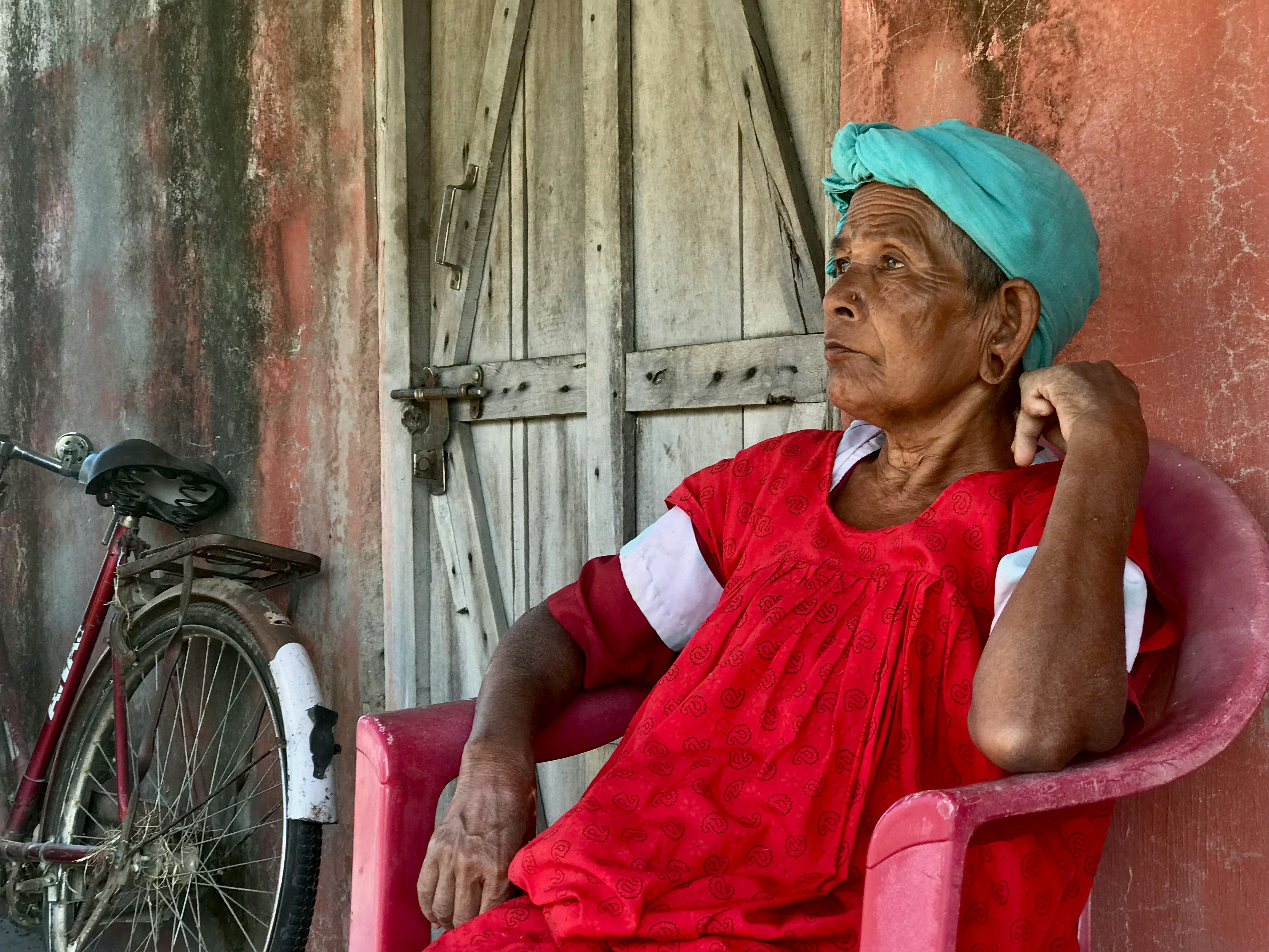 Elderly woman in red dress wearing turquoise headscarf sits.