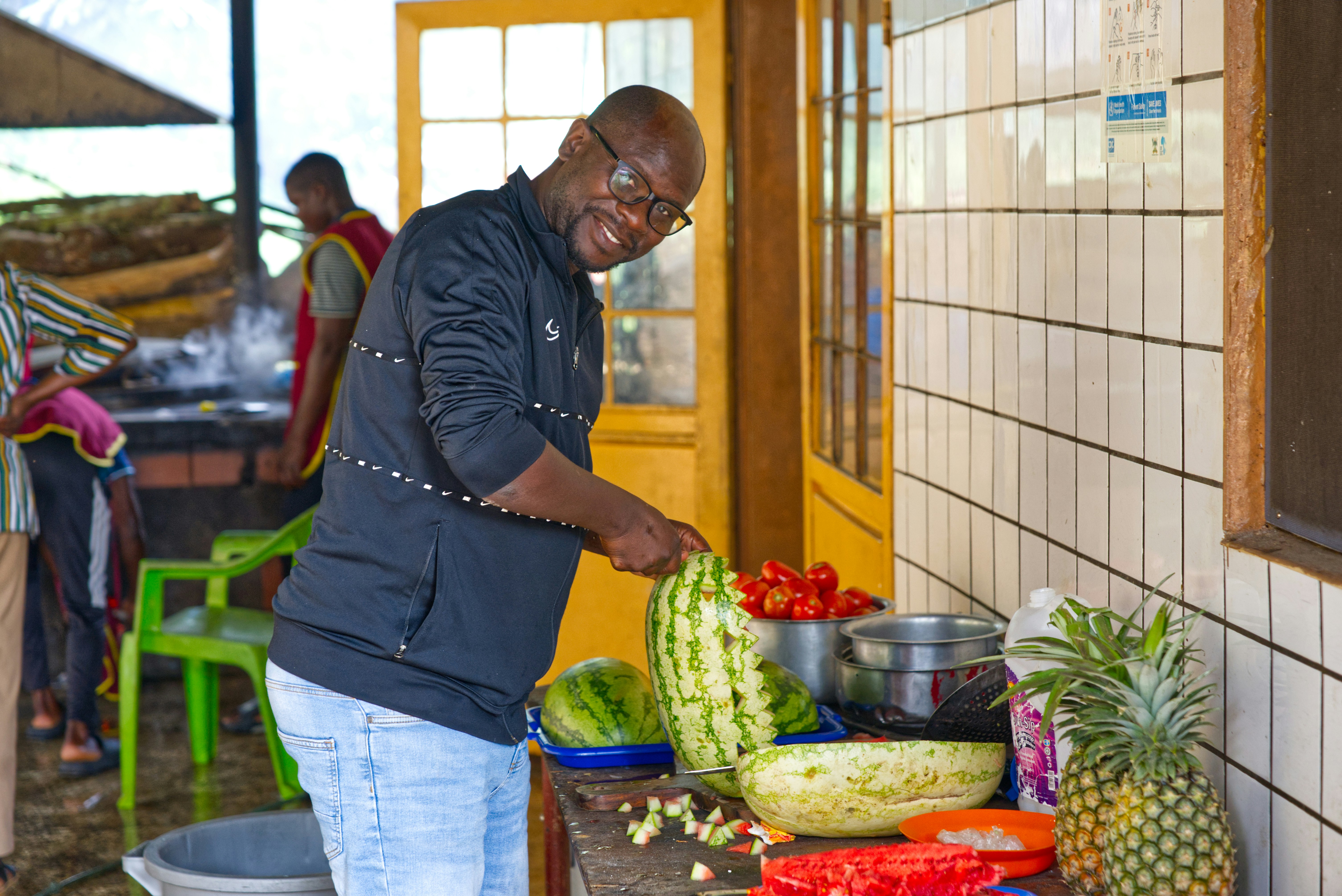 Man slicing watermelon in a kitchen with fruit.