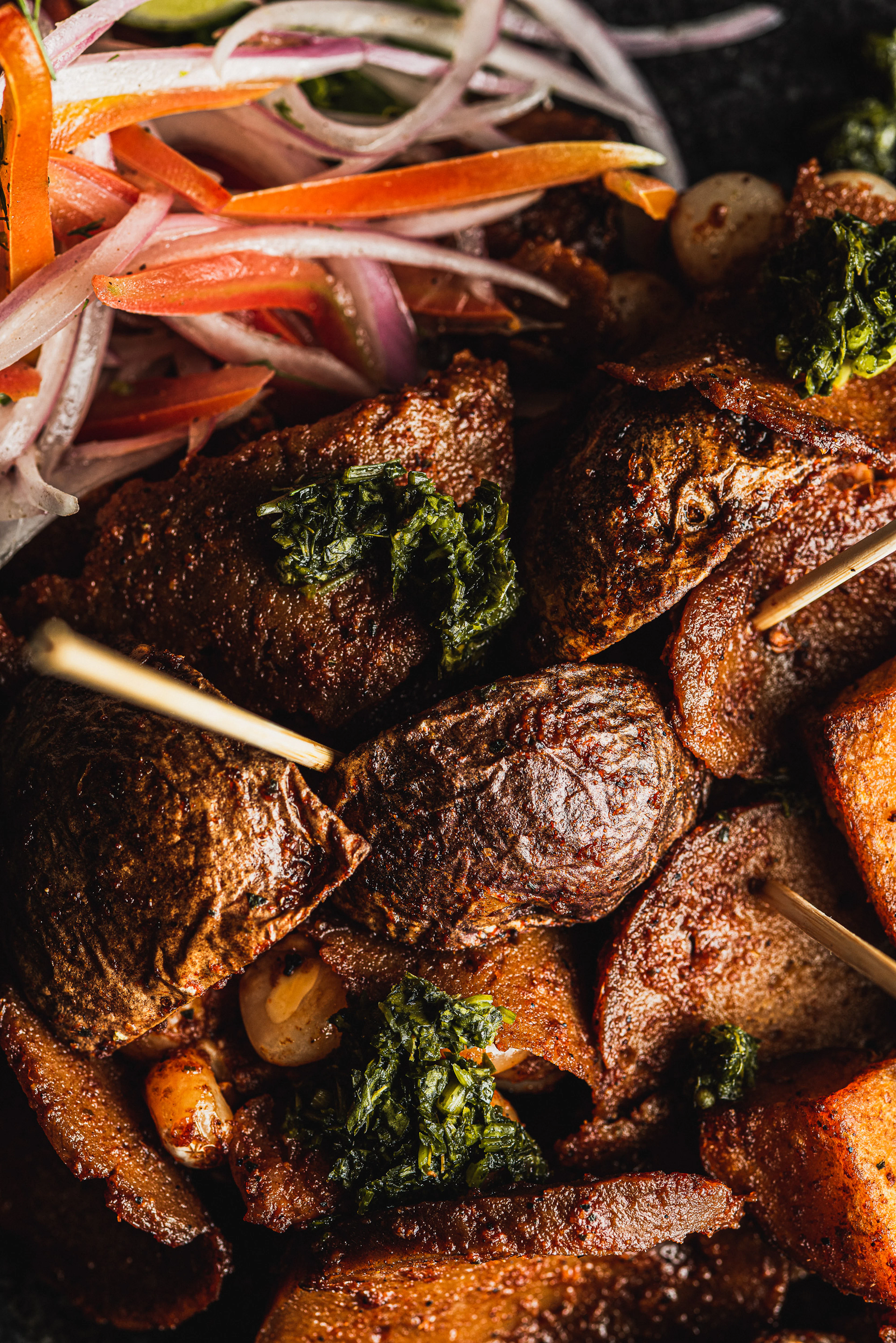 A close-up of Peruvian anticuchos — charred skewered beef heart glossy with marinade, dressed with green huacatay sauce and heaped with salsa criolla of sliced red onion and tomato, alongside golden potatoes and choclo corn kernels