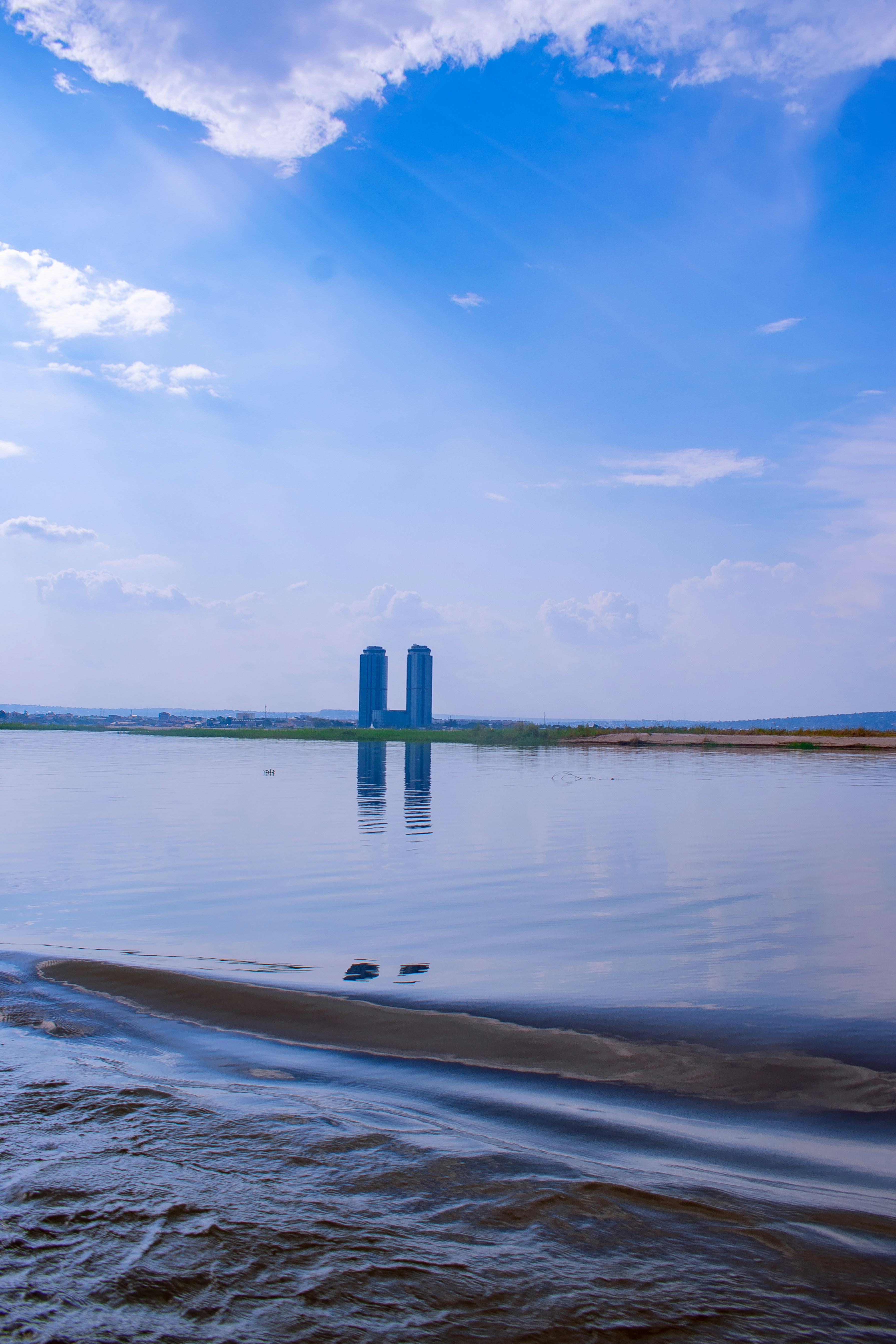 Two tall buildings reflected in calm water under blue sky.