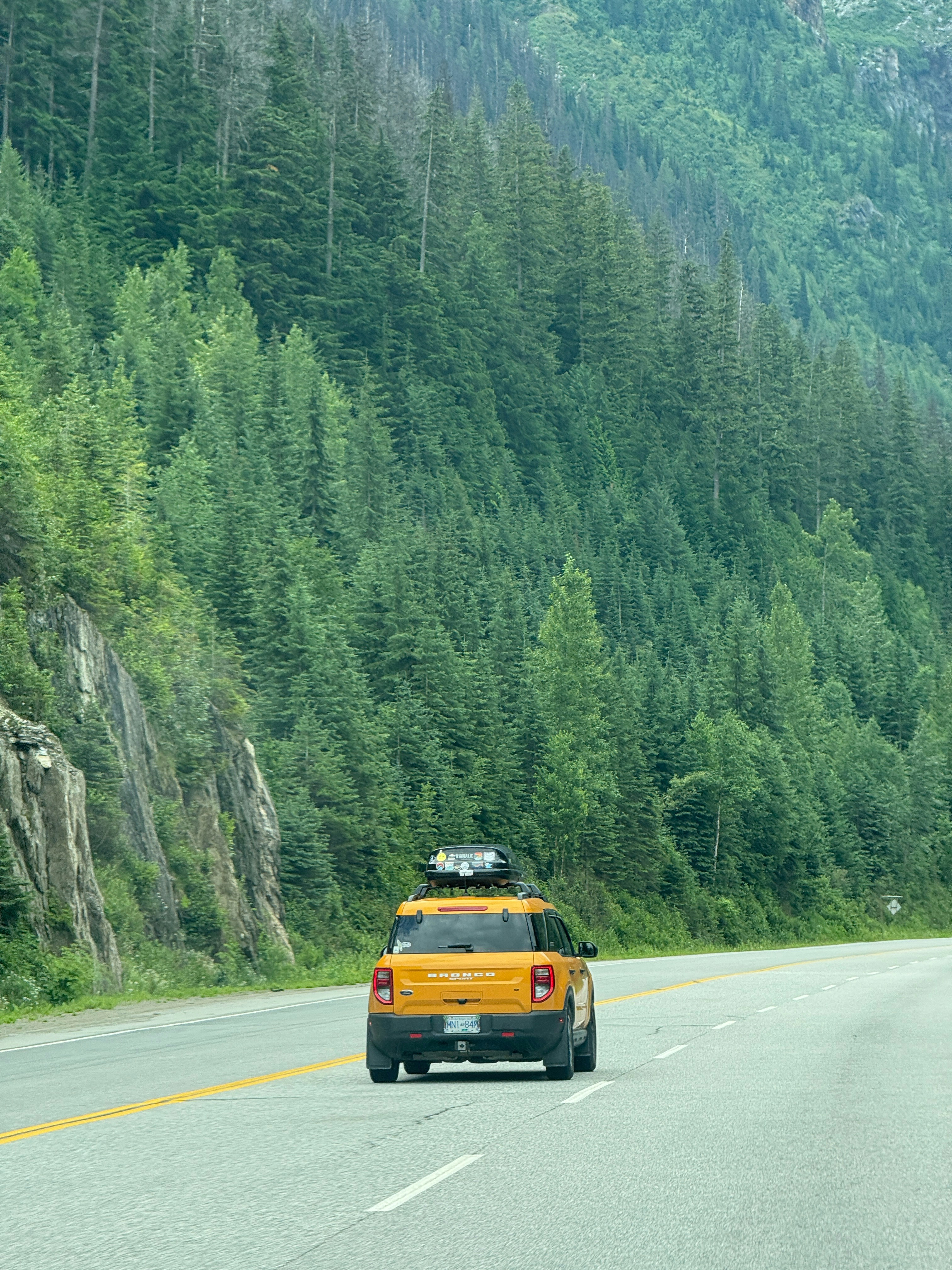 Yellow car drives on highway beside forested mountain.