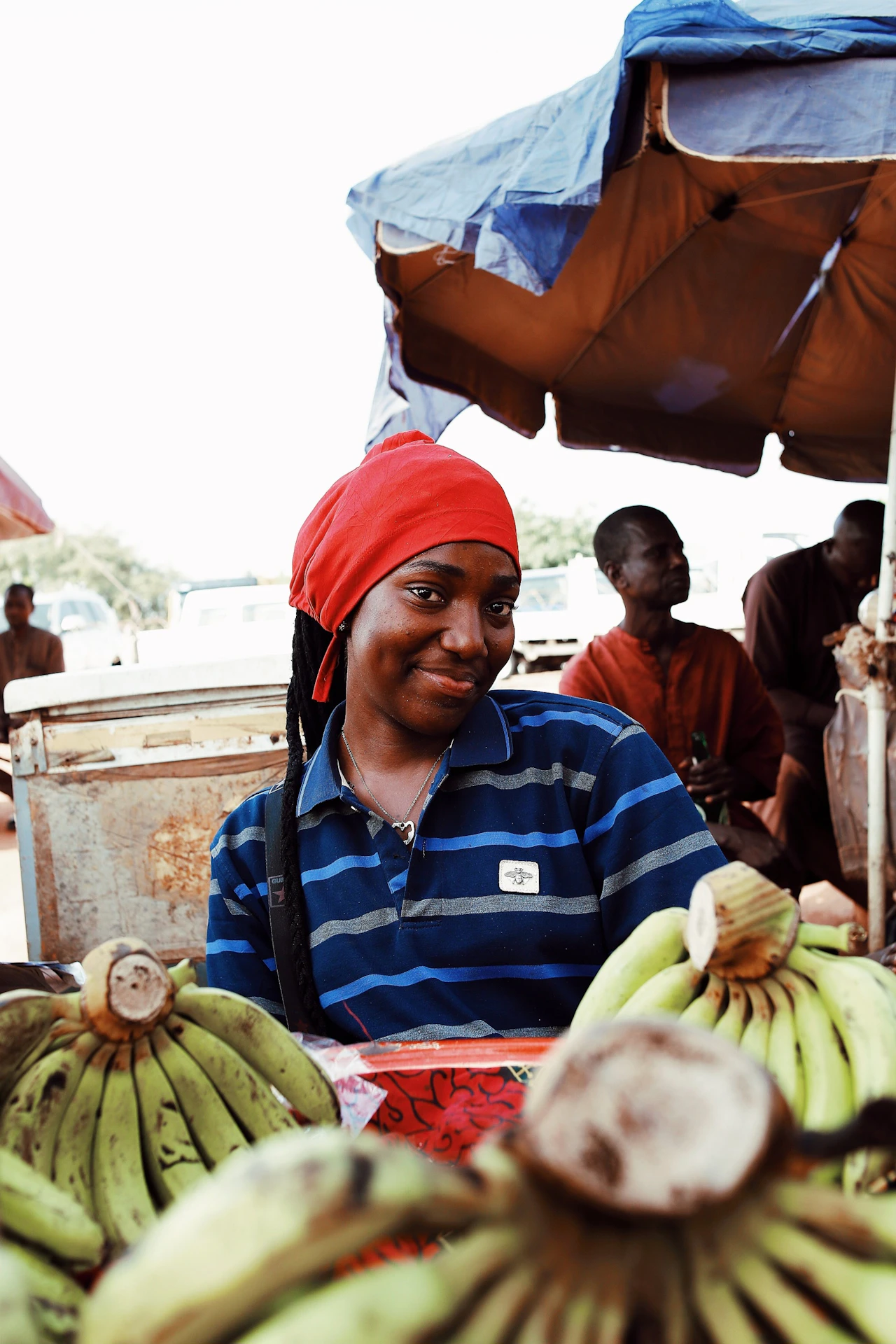Stallholder in a red headscarf smiles behind a pile of green bananas at an outdoor market.