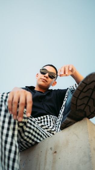 Man in sunglasses and checkered pants sits on concrete.