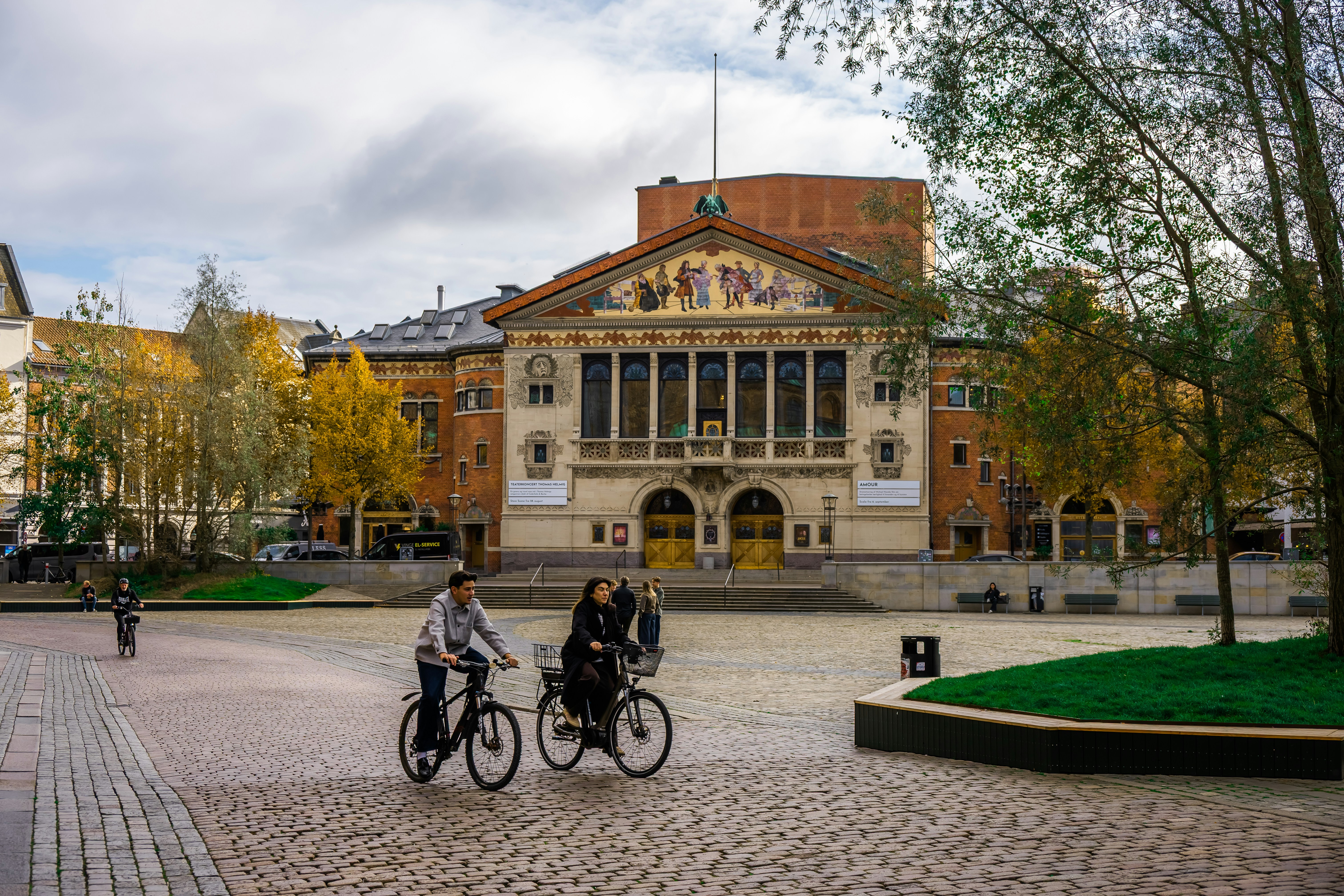 Two people cycle past a grand building in autumn.