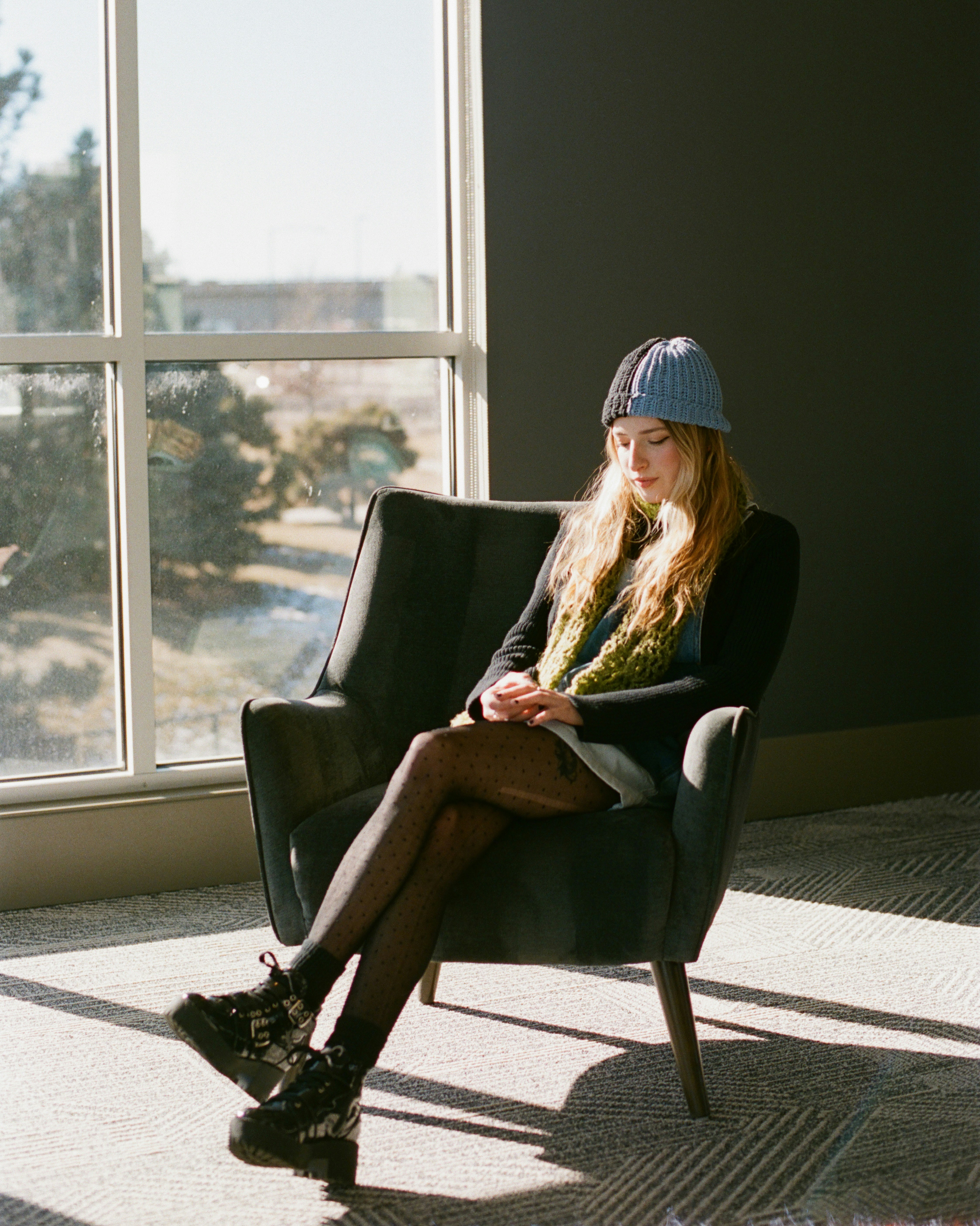 Young woman in hat sits in armchair by window.