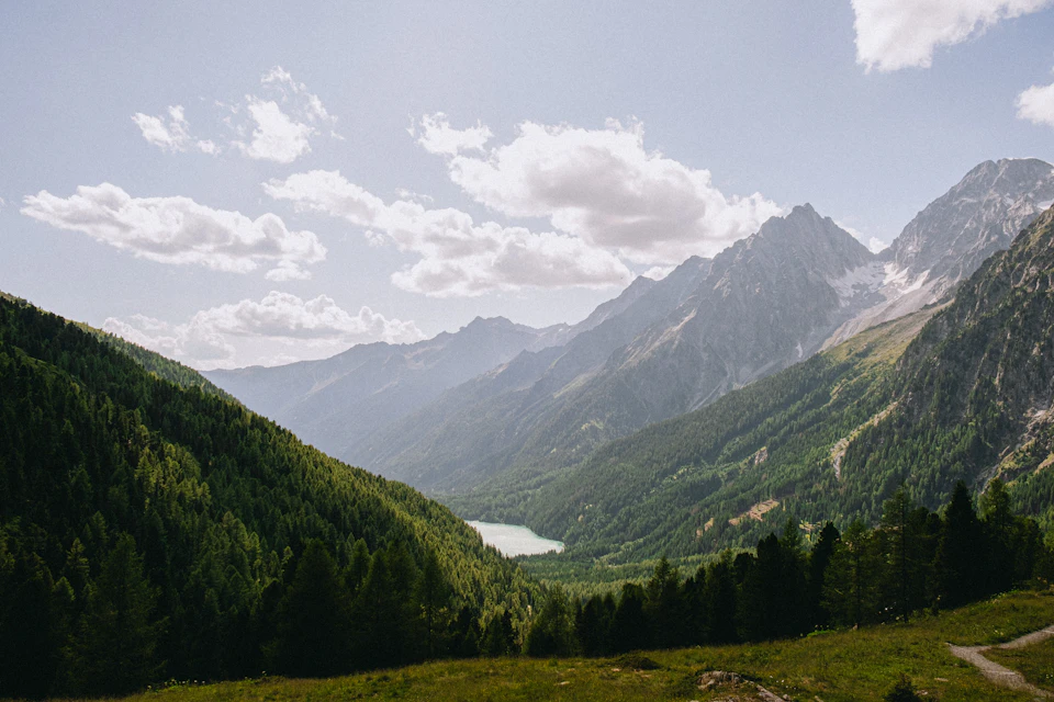 The image depicts a breathtaking view of a valley nestled between two majestic mountains, with a lake at its center. The valley is lush and green, dotted with trees that add depth to the scene. The mountains, covered in dense forests, rise high into the sky, their peaks touching the clear blue expanse above. The sky above is a beautiful shade of blue, lightly speckled with clouds. The perspective of the image is from a higher vantage point, looking down upon the valley and its surroundings, creating a sense of awe-inspiring grandeur.