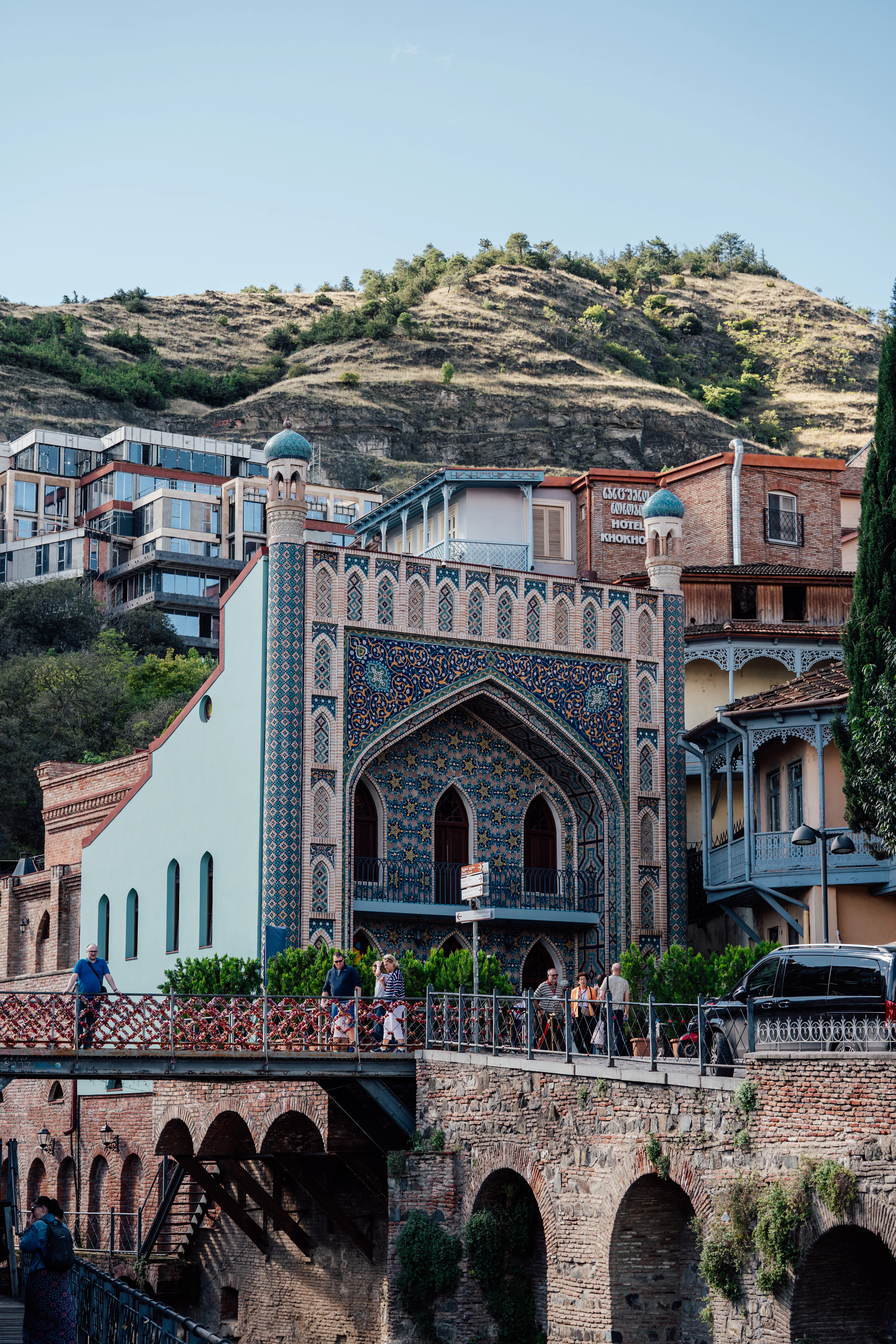 The blue-and-terracotta tiled Persian facade of the Orbeliani sulphur bathhouse in Tbilisi's Abanotubani quarter, above brick vaults and a walkway, with the bare hillside rising behind