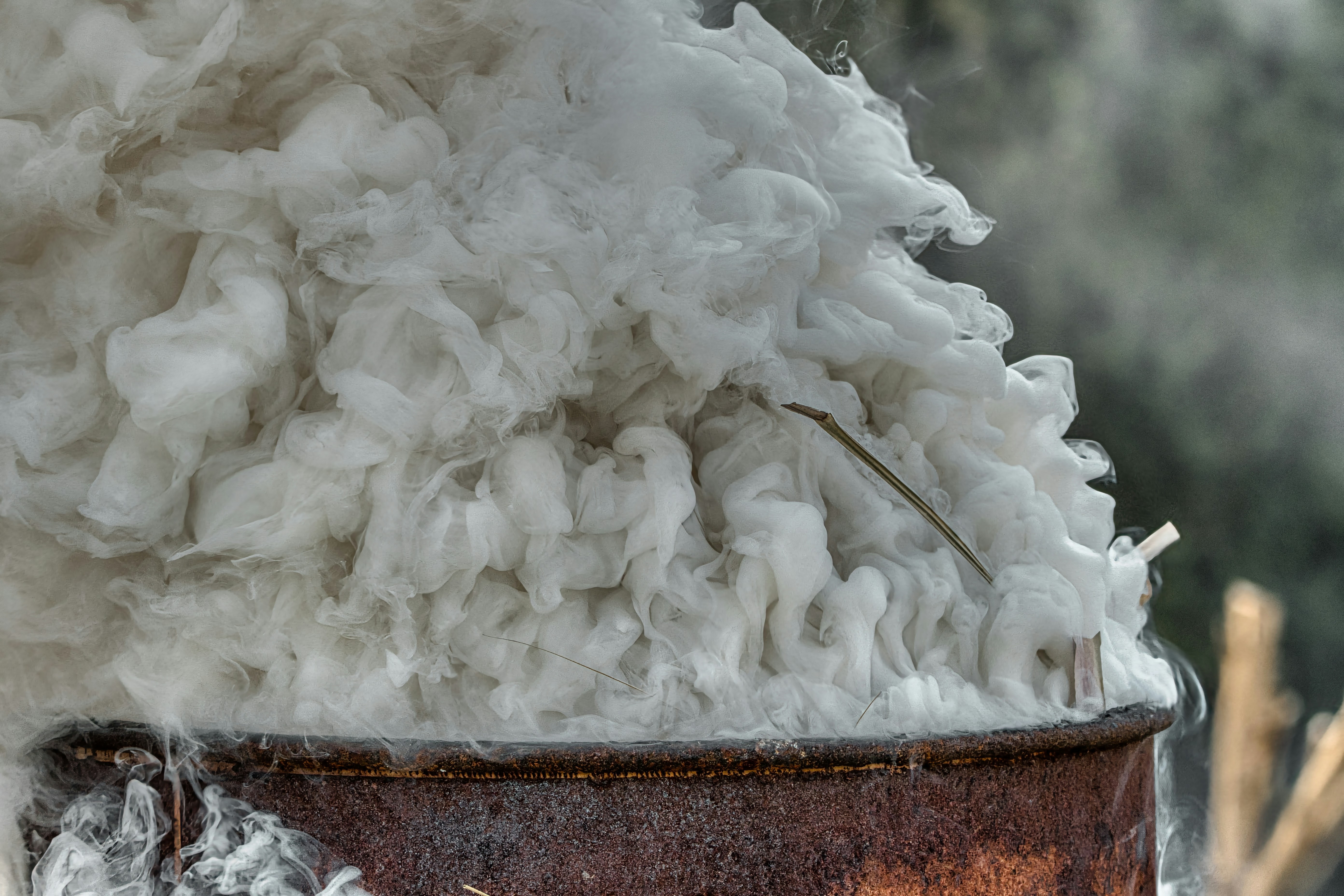 White smoke billows from a rusty barrel outdoors.