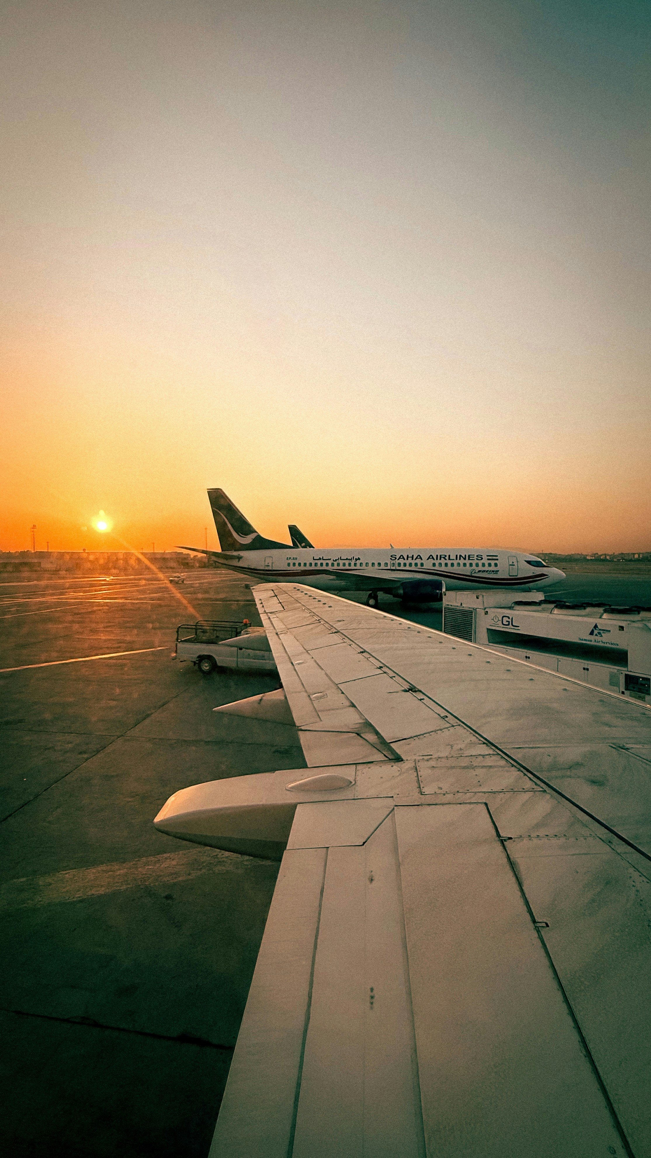 Airplanes on tarmac at sunset with wing in foreground.